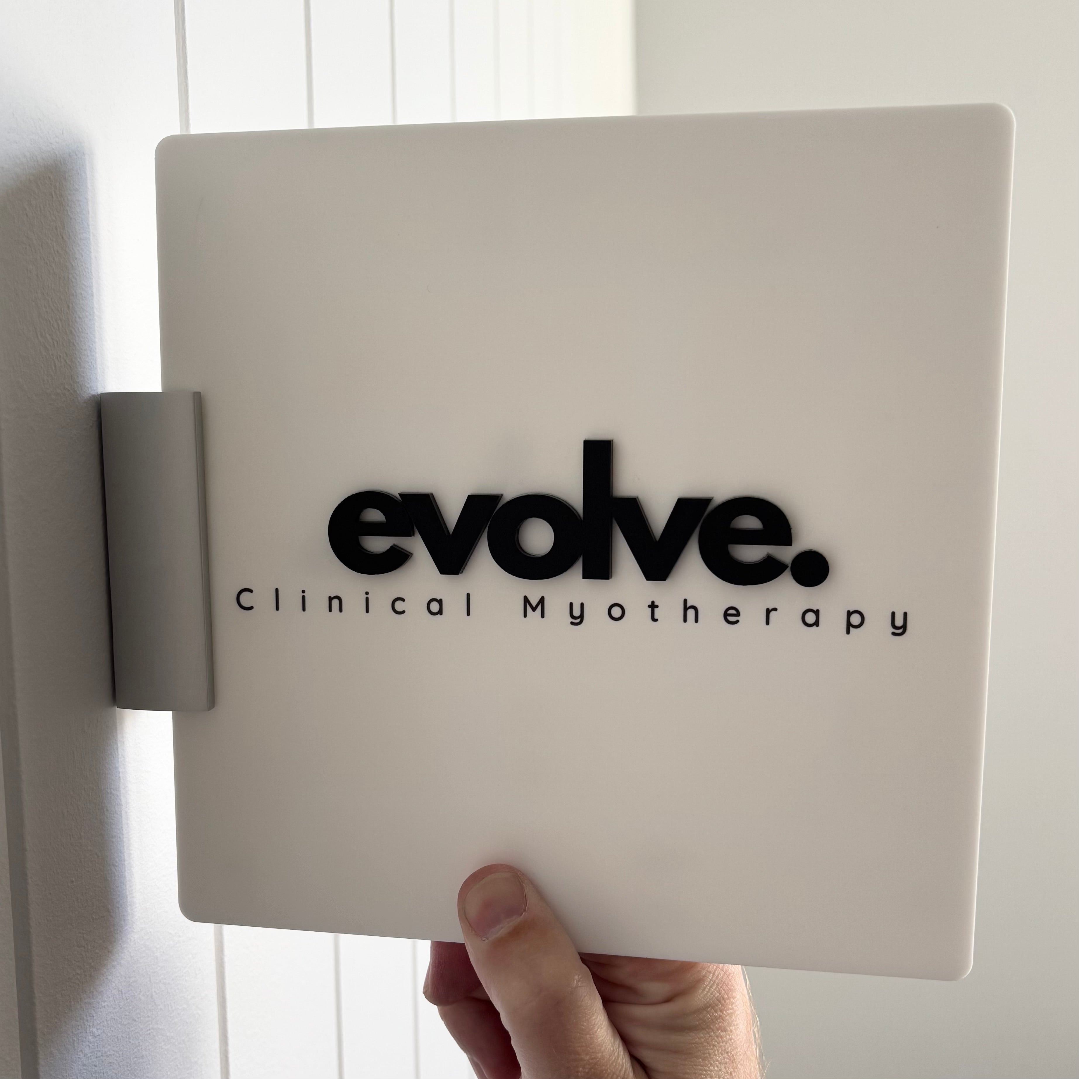 A hand holds a square white corridor sign with black raised lettering that reads “evolve. Clinical Myotherapy.” The sign is mounted on a silver side bracket, designed to project from a wall in a hallway.