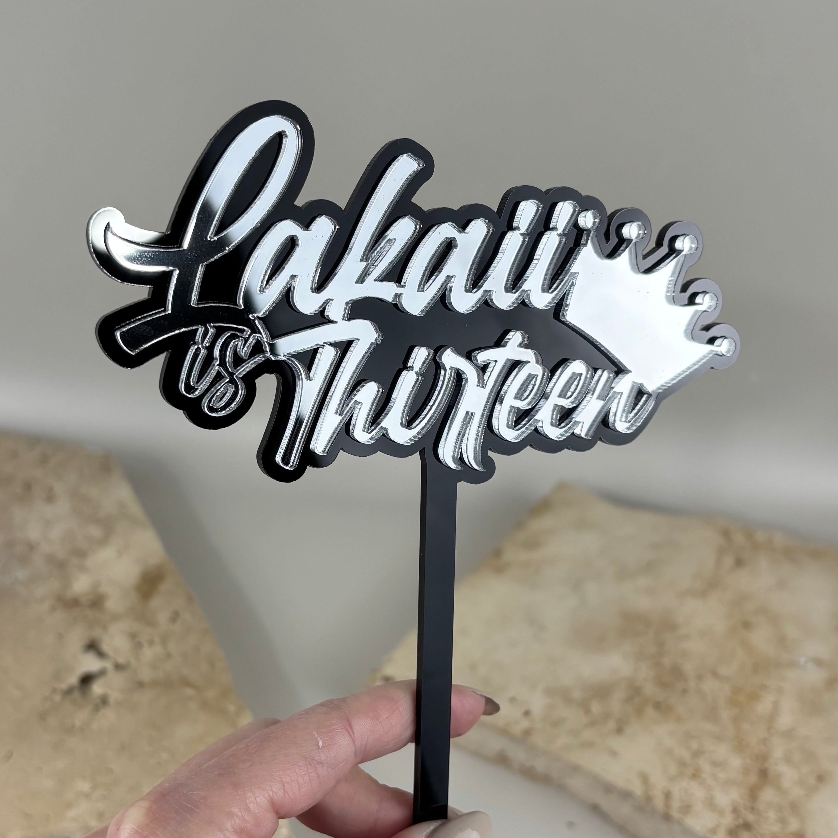 Custom black and silver mirror acrylic 3D cake topper with personalised Lakaii is Thirteen text and crown design for a 13th birthday.