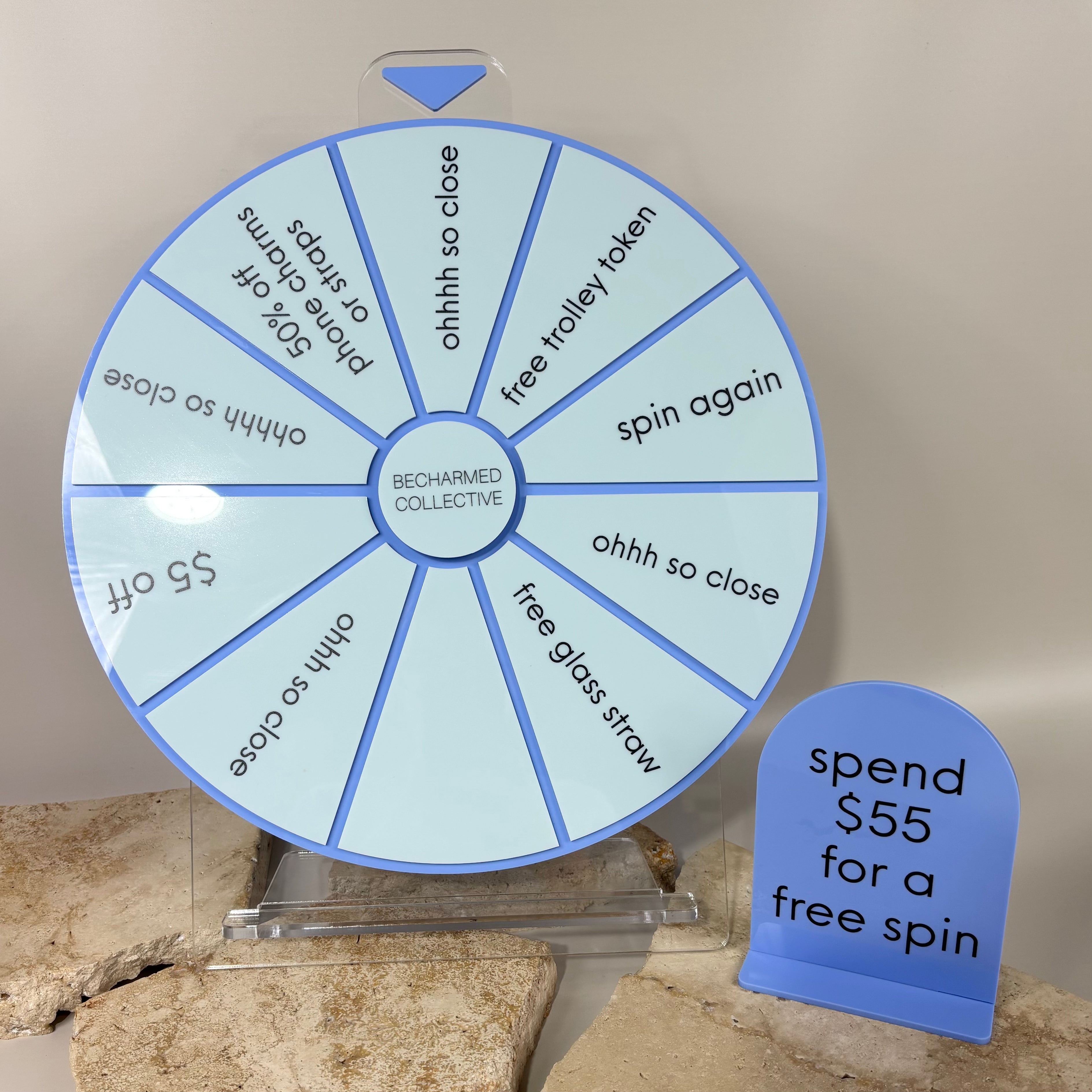 Custom blue spin the wheel prize display for Becharmed Collective, featuring rewards like discounts, free gifts, and extra spins, with a sign reading ‘spend $55 for a free spin.
