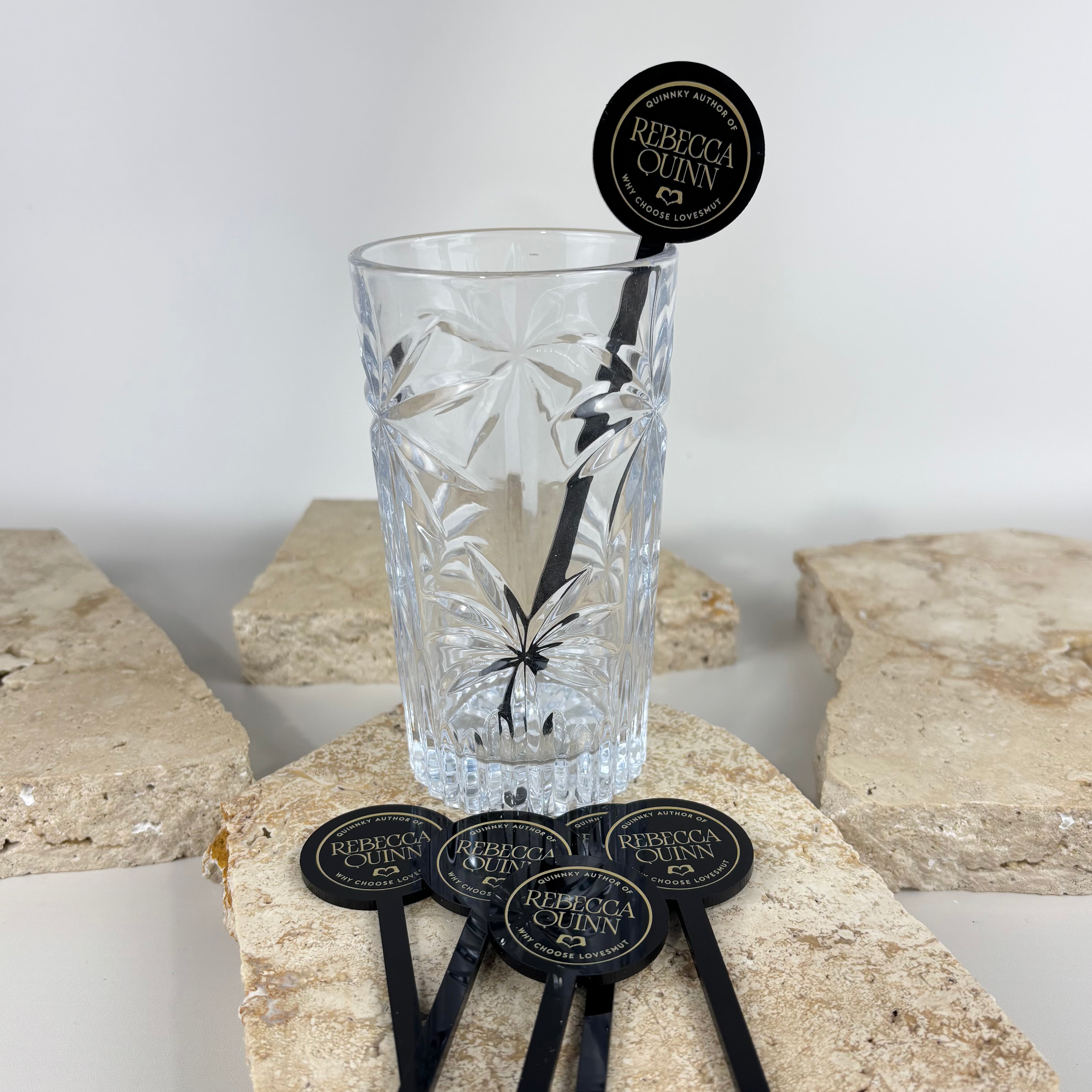 Custom black acrylic drink stirrers with round tops printed with “Rebecca Quinn” and “We Choose Love” branding, displayed with a crystal glass on textured stone slabs.