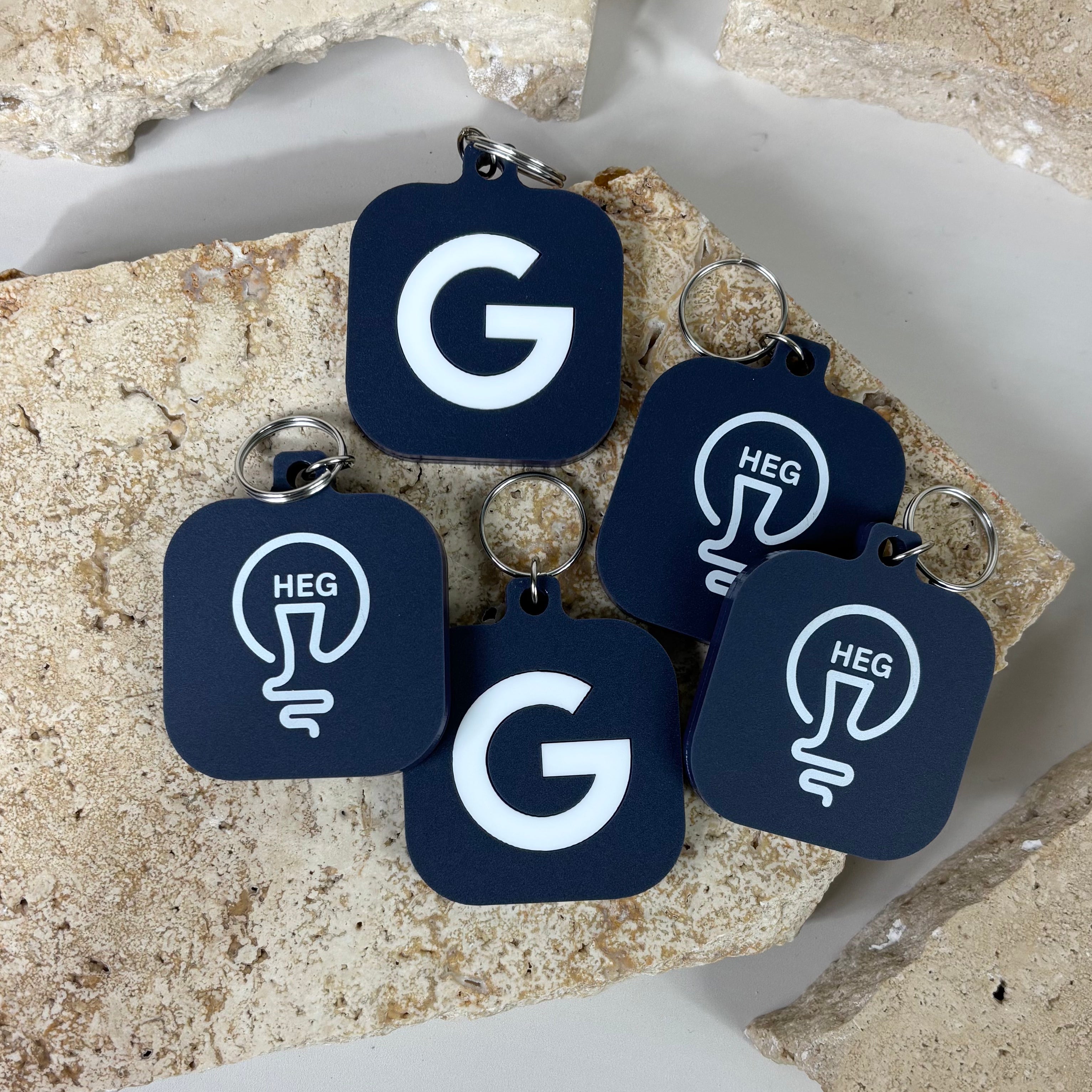 Navy blue square NFC keychains with rounded corners displayed on textured stone tiles. Some keychains show a bold white letter G and others show a white lightbulb design with the letters HEG inside. Each NFC keychain has a silver keyring and is designed for digital business cards or smart contact sharing.