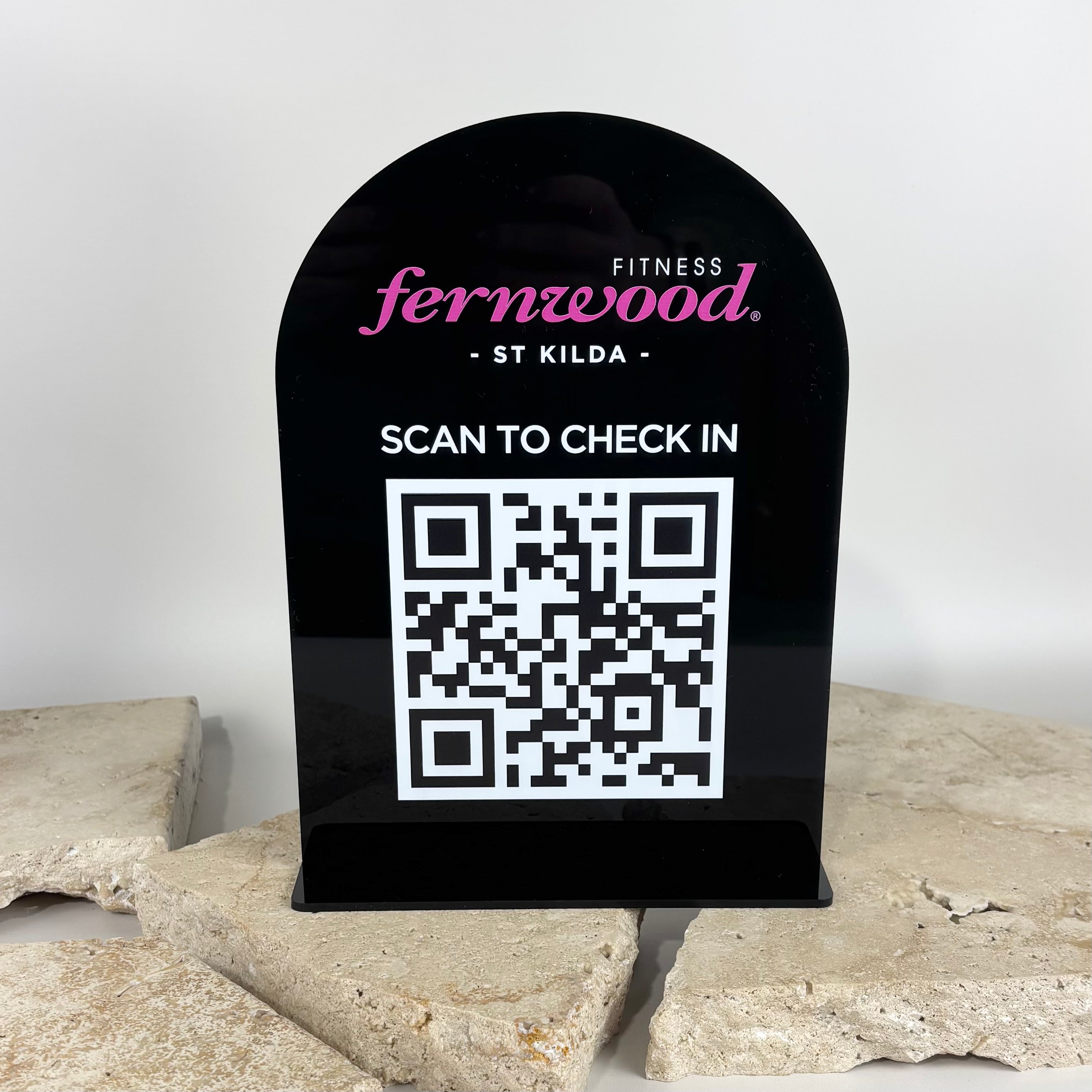 Black acrylic arched counter stand with Fernwood Fitness St Kilda logo, white ‘Scan to Check In’ text, and large QR code for customer check-in, displayed on stone tile background