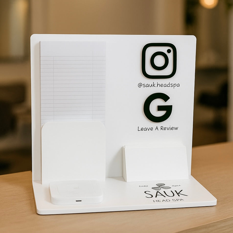 Custom white salon brochure holder displayed on a wooden reception desk, featuring spaces for booking sheets, business cards, a payment terminal, and social media icons for Instagram and Google Reviews, with the SAUK Head Spa logo printed on the base.