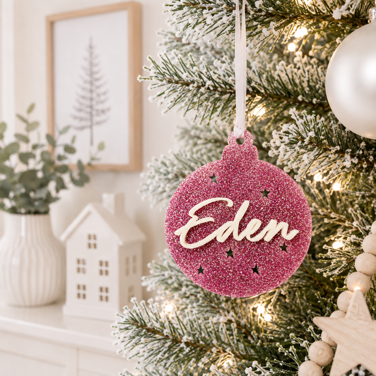 Personalised pink glitter acrylic Christmas bauble with custom 3D name and star cutouts, hanging on a modern frosted Christmas tree.