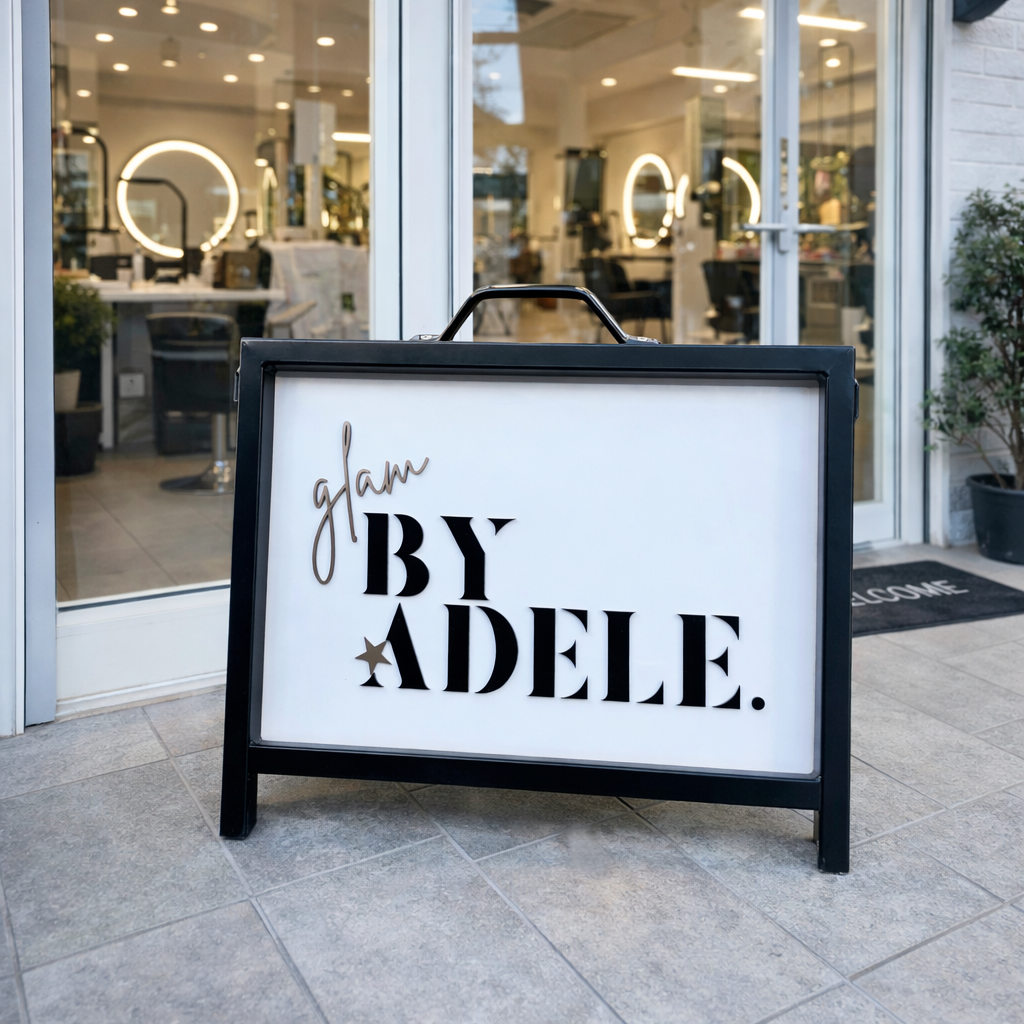 Black a frame salon sign placed outside a hair and beauty salon entrance, featuring custom acrylic lettering with “glam by Adele” branding on a white panel