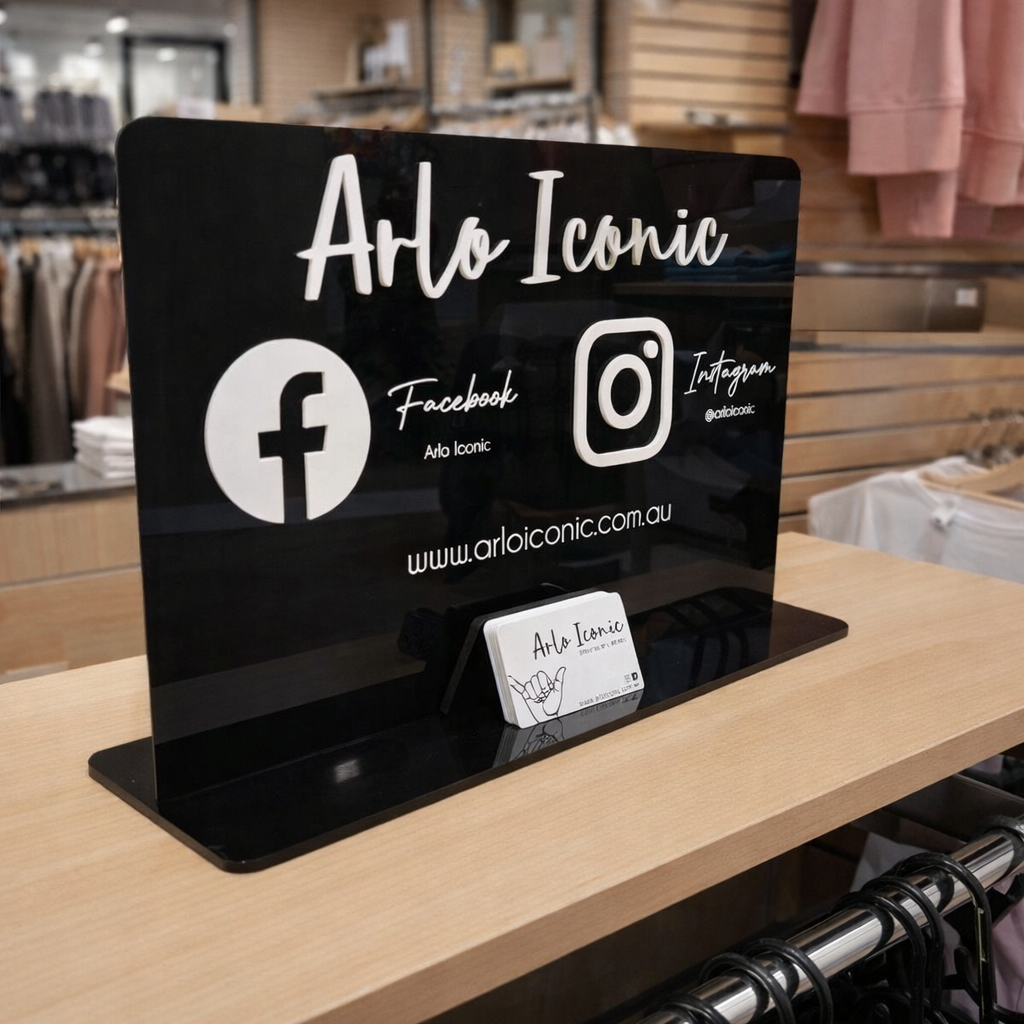 Black acrylic social media business stand displayed on a clothing store counter, featuring Facebook and Instagram icons, website URL and business card holder, custom retail signage designed for fashion boutiques and clothing stores in Australia