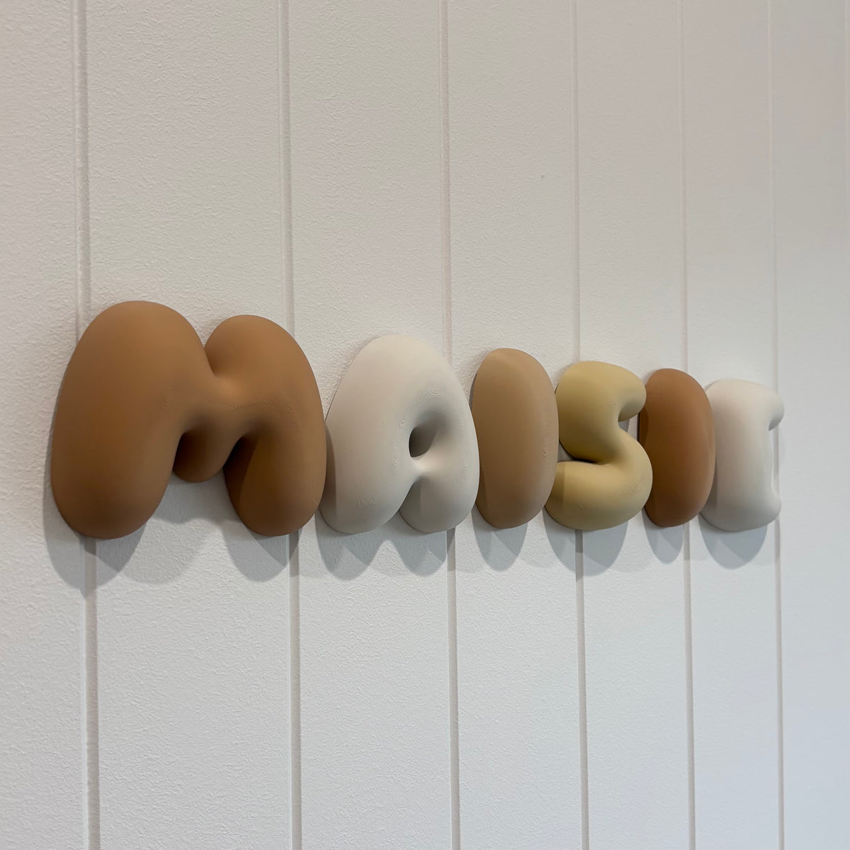 Custom 3D bubble name wall sign spelling “Maisie” in neutral earthy colours, mounted on a white panelled wall.