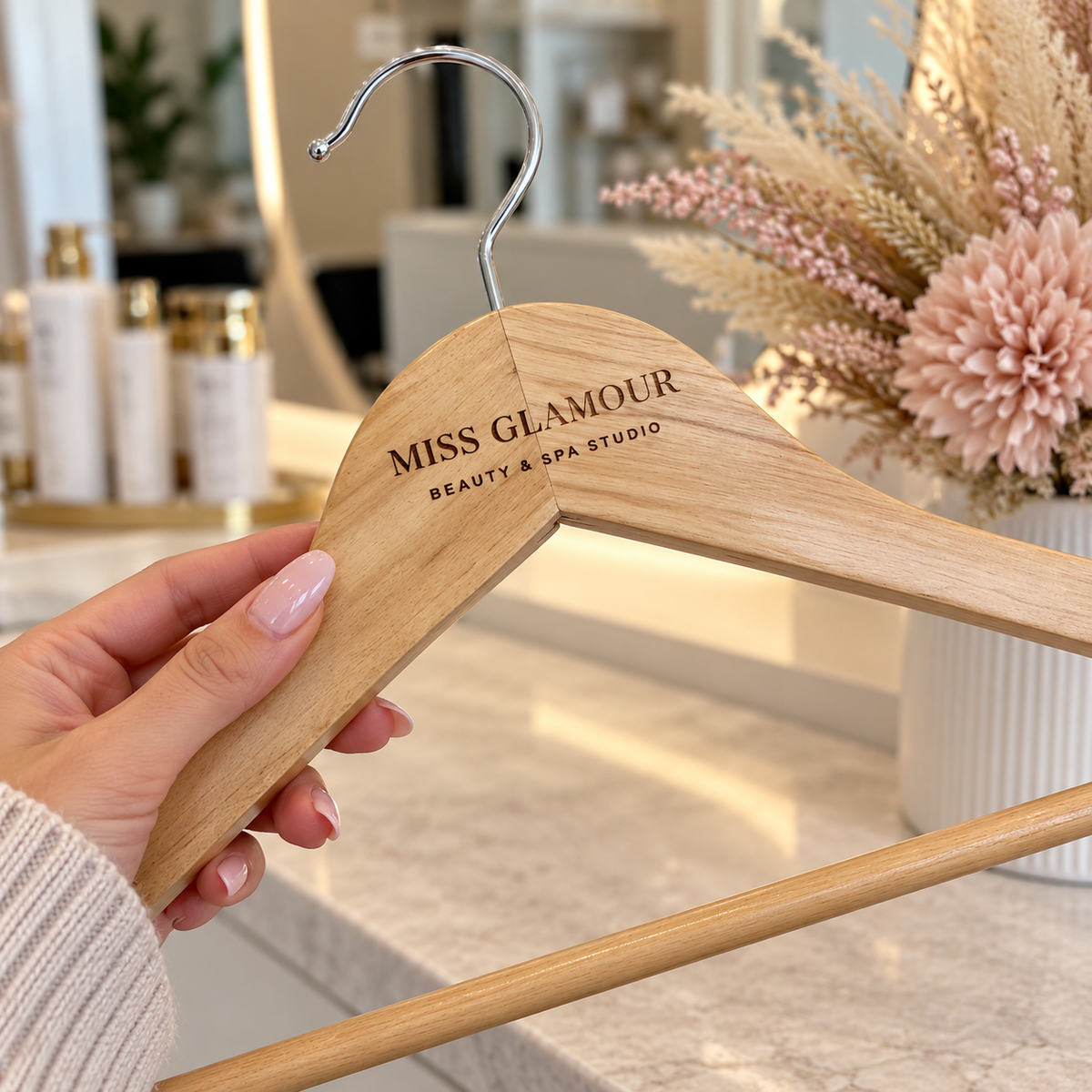 Custom engraved wooden coat hanger with Miss Glamour Beauty & Spa Studio logo, held by a hand with pink manicured nails in a modern beauty salon setting.