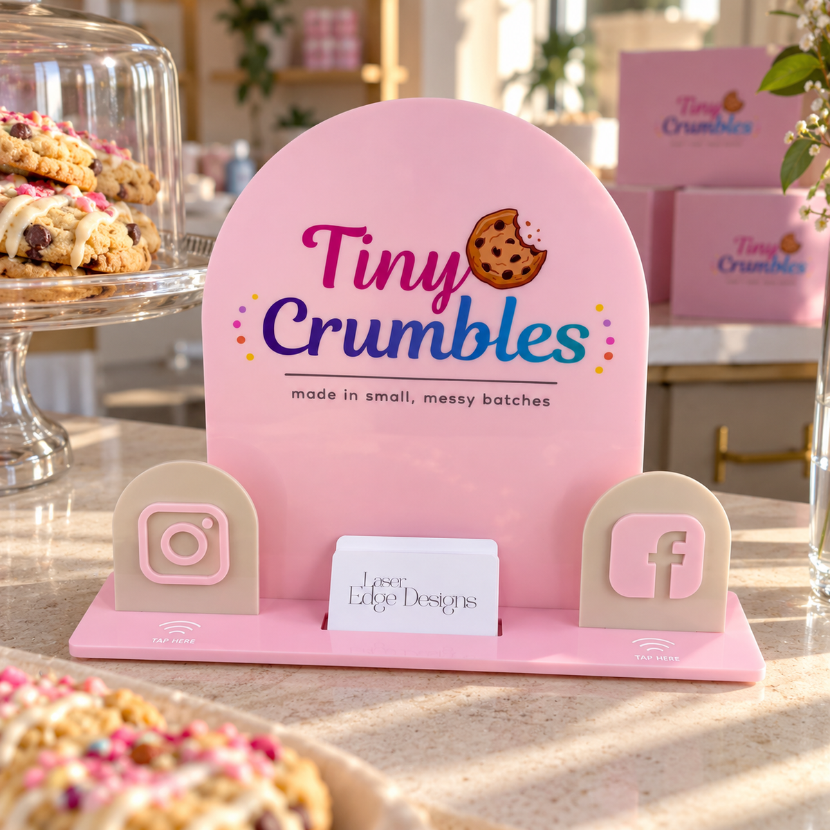 Custom pink acrylic NFC business display for a cookie and buttercream cake business, featuring Instagram and Facebook tap-to-connect icons and a business card holder on a bakery counter.