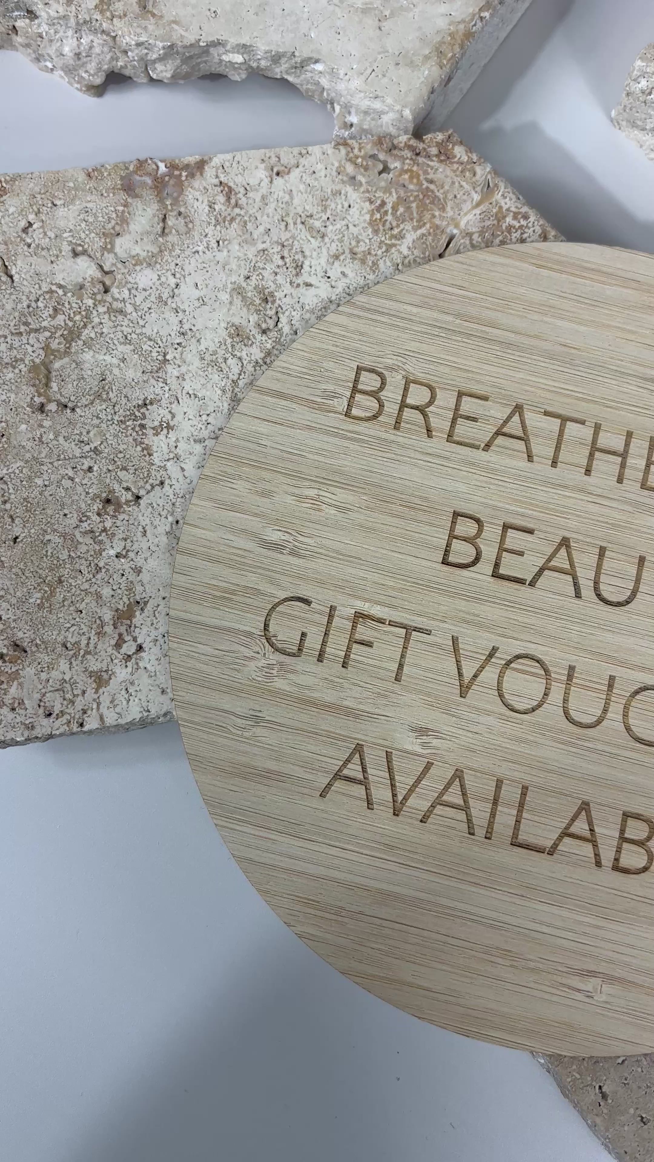 Round engraved bamboo sign reading ‘Breathe Easy Beauty Gift Vouchers Available’, displayed on stone background.