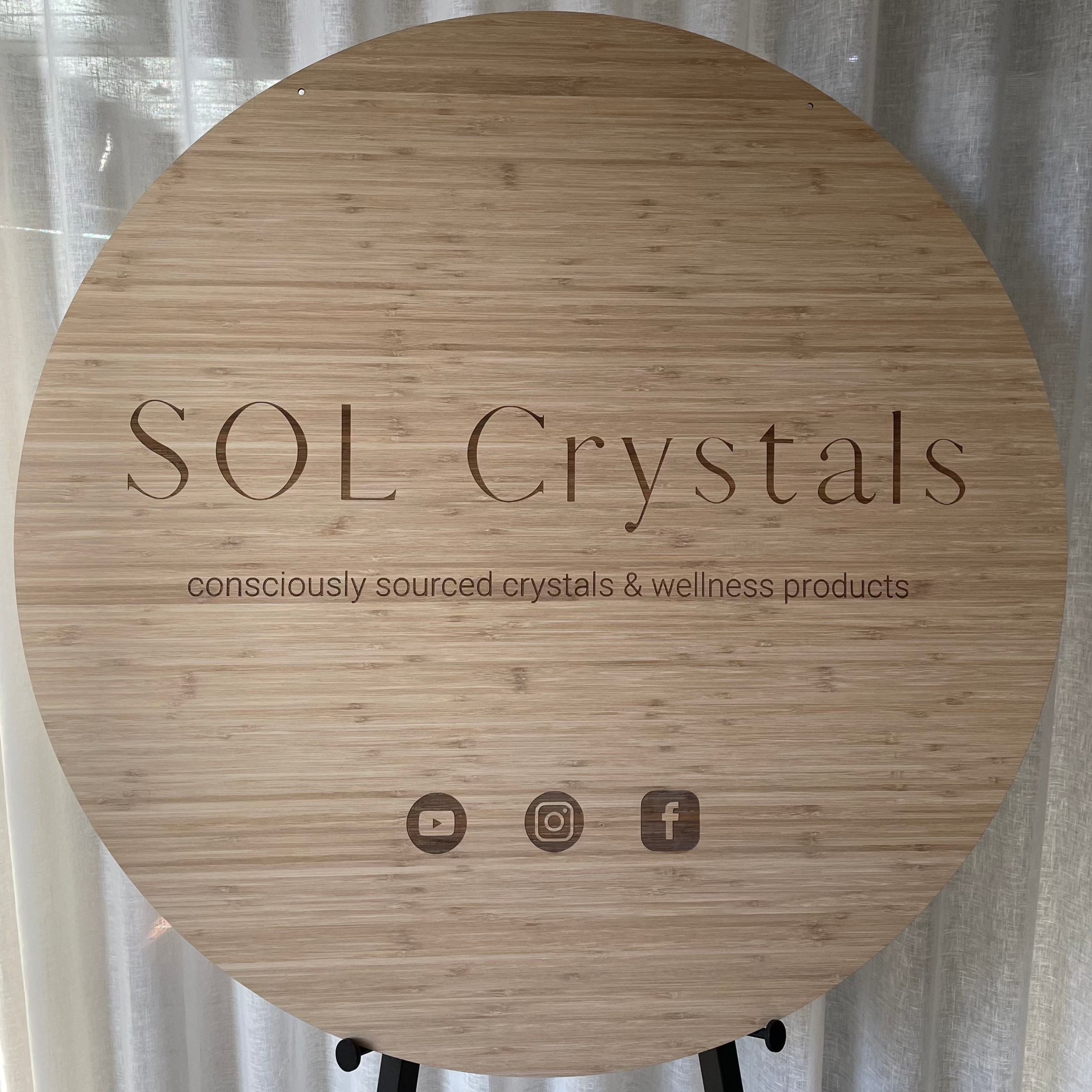 SOL Crystals, Round bamboo sign with engraved minimalist logo and social icons, displayed against neutral curtains.