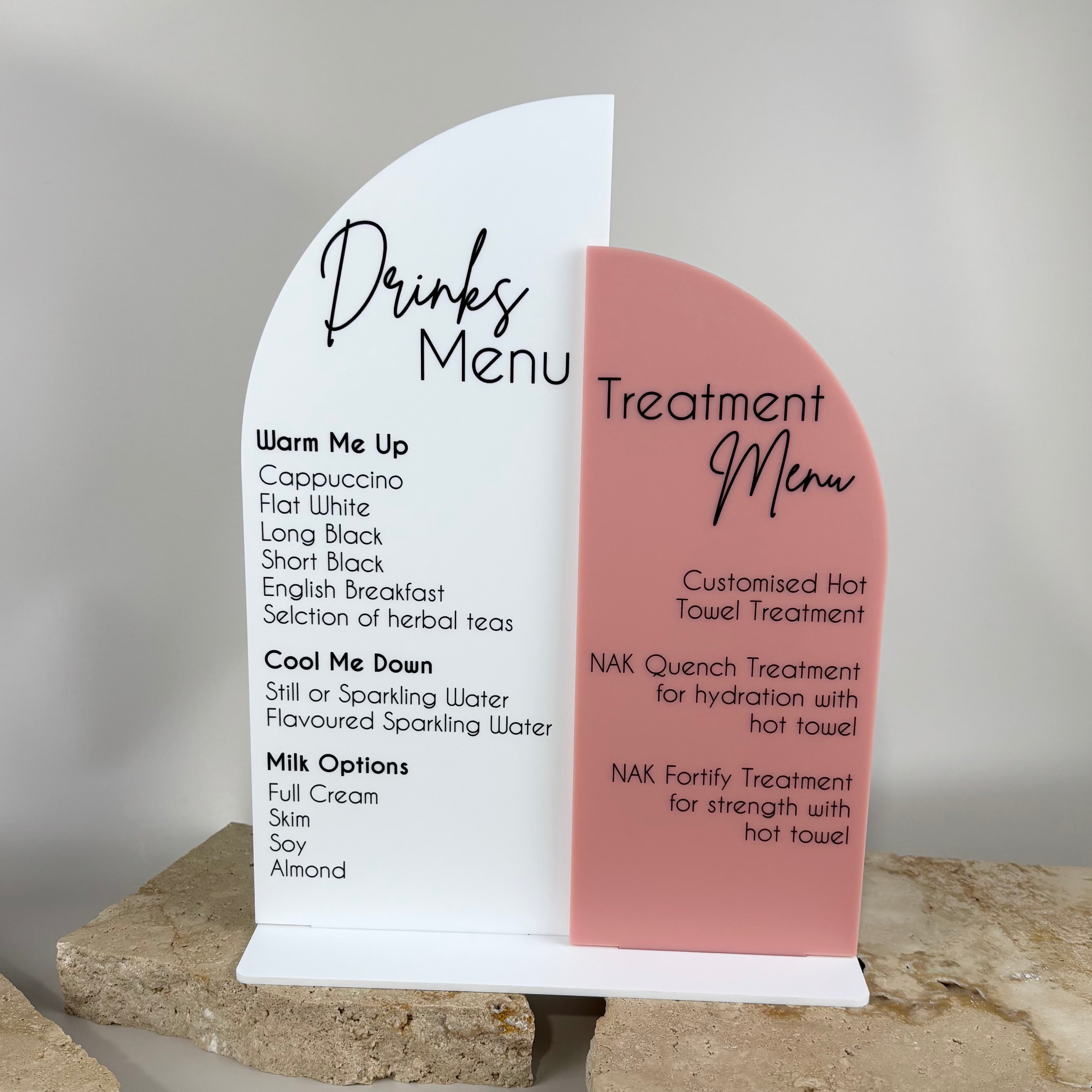 Modern acrylic salon counter sign featuring a white and blush pink arch design with a printed drinks menu and treatment menu, listing coffee options, teas, sparkling water, and NAK hot towel treatments, displayed on stone slabs.