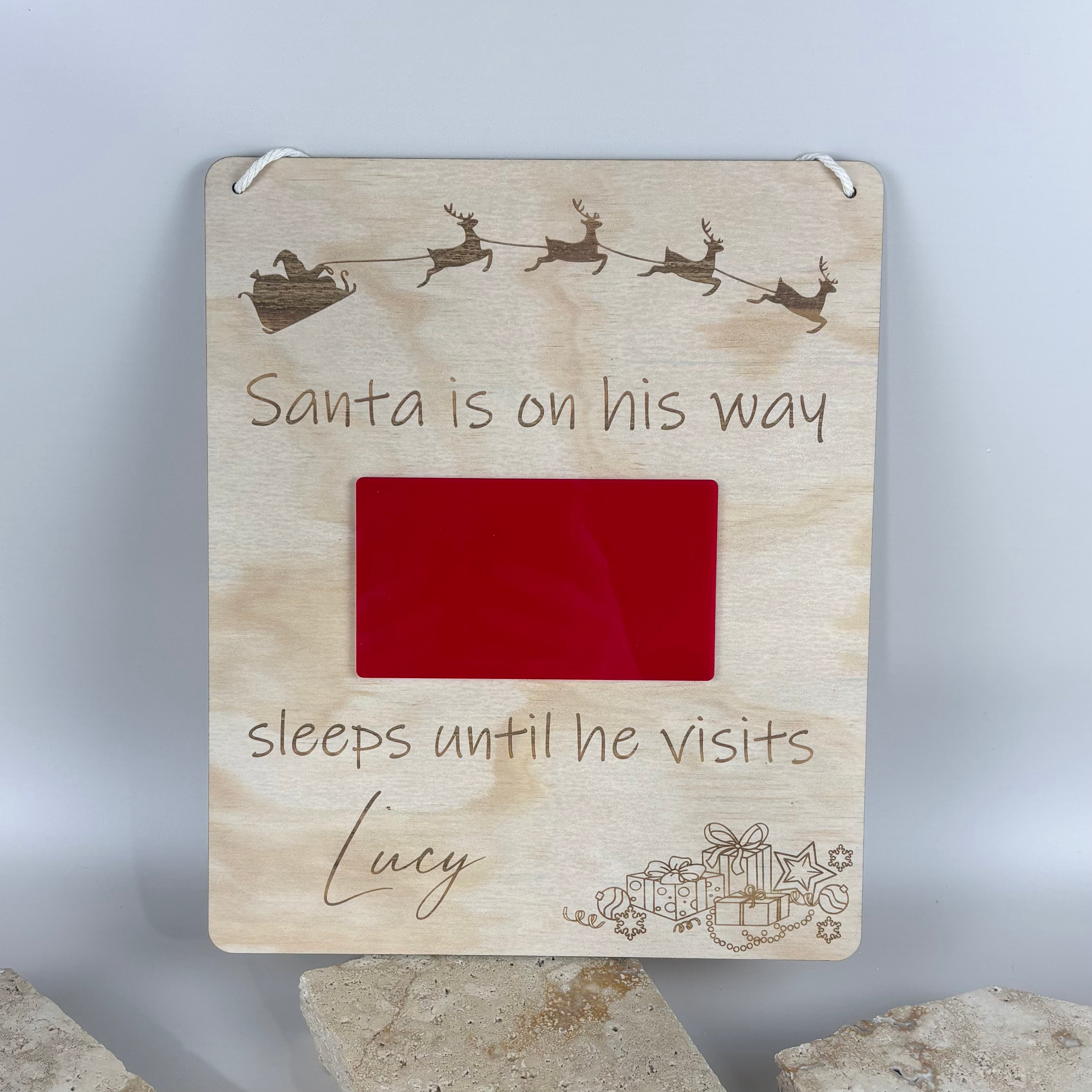 Personalised wooden Christmas countdown sign with engraved Santa sleigh design, red acrylic countdown window and custom child’s name “Lucy,” engraved text reading “Santa is on his way, sleeps until he visits,” displayed on stone backdrop.