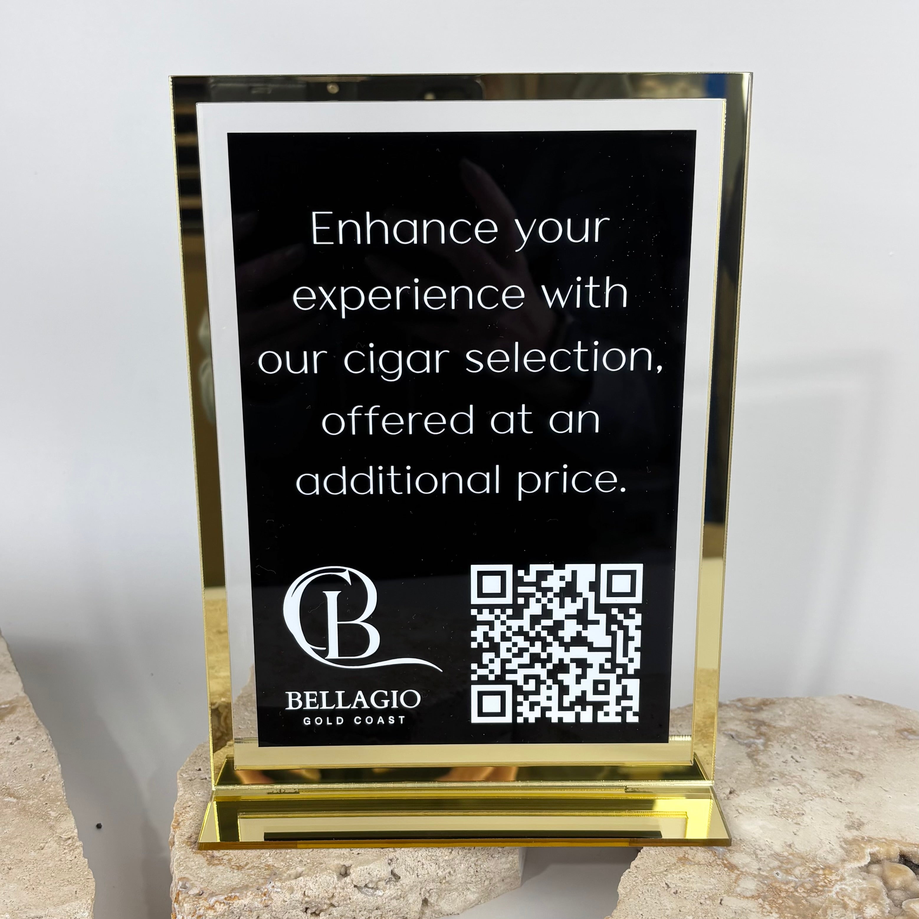 Gold acrylic counter sign displaying a black insert with white text promoting a cigar selection, featuring a QR code and the Bellagio Gold Coast logo, styled on a stone surface in a studio setting.