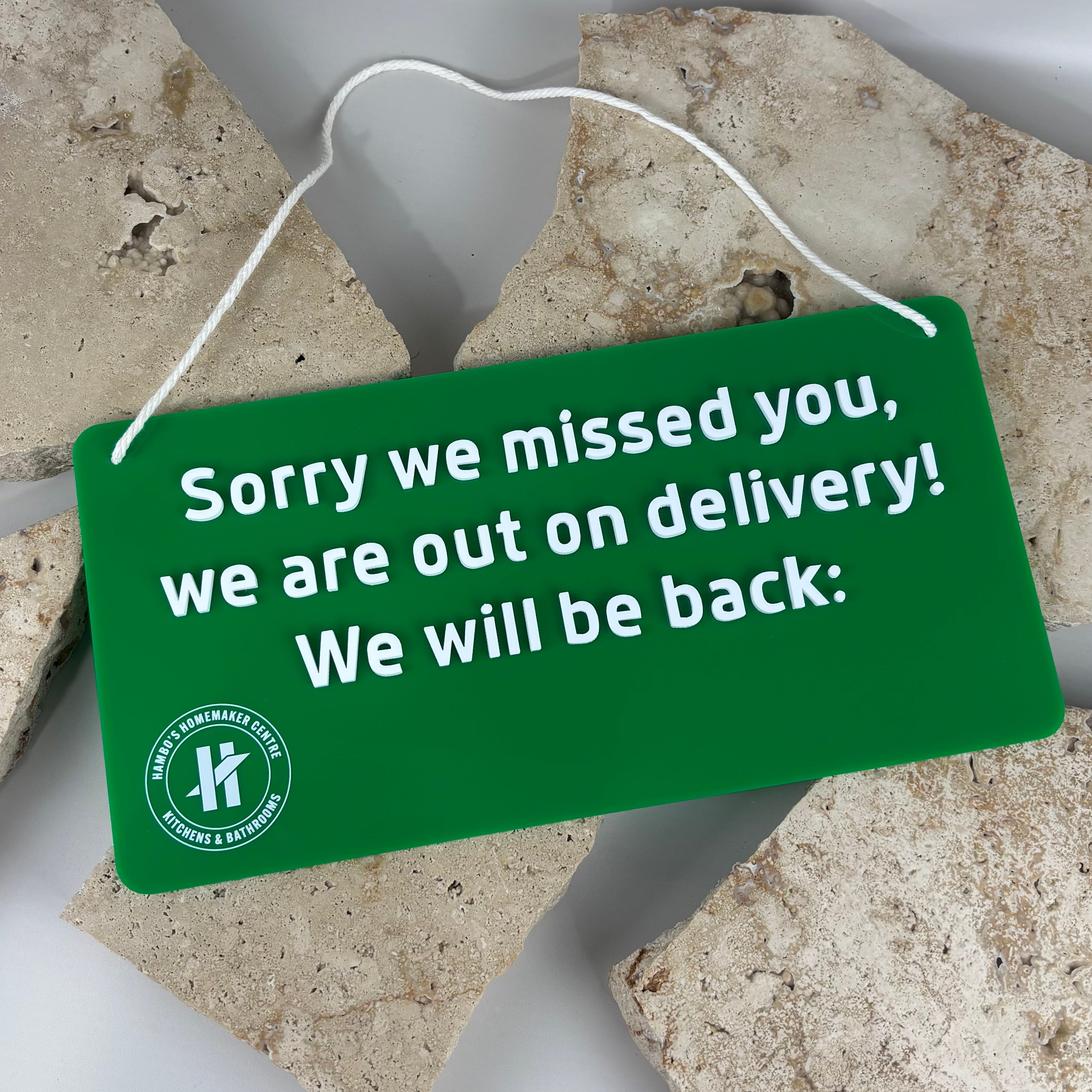 Green acrylic hanging sign with white raised text reading “Sorry we missed you, we are out on delivery. We will be back.” featuring rope handle and Hambos Homemaker Centre logo, styled on a neutral stone background