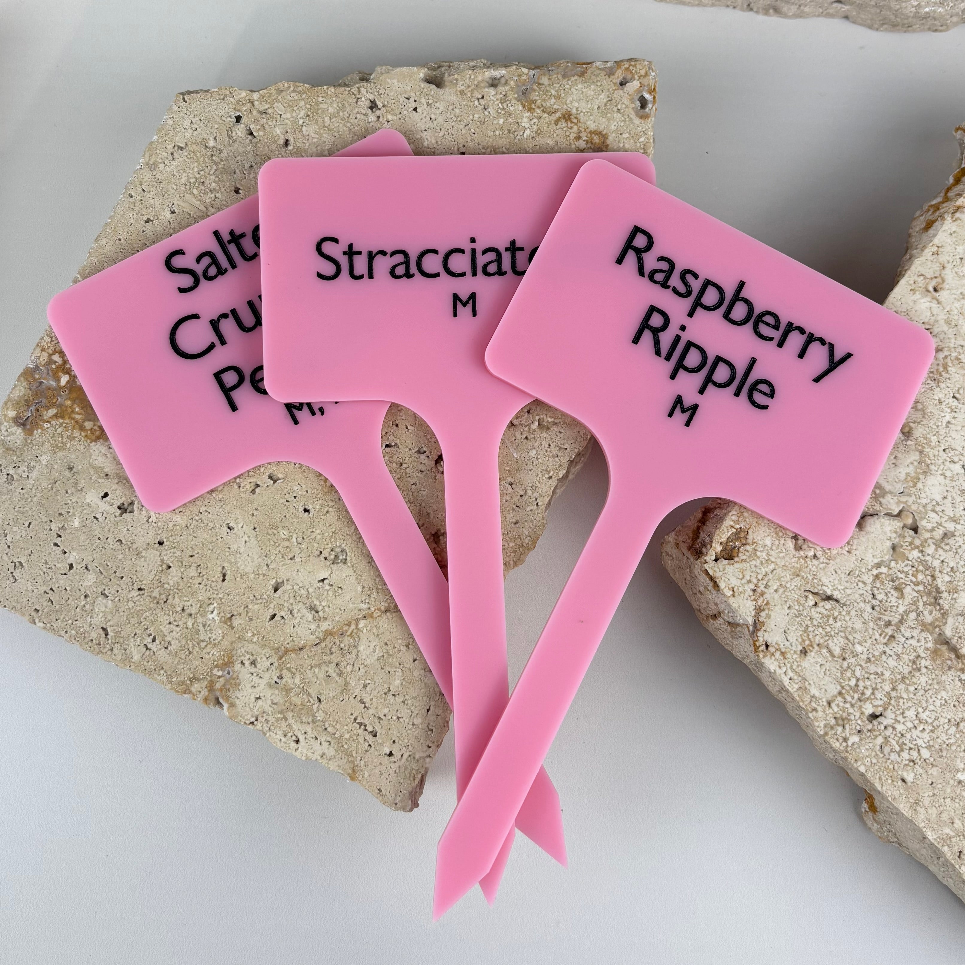 Pink acrylic ice cream flavour sticks with black engraved text reading Raspberry Ripple, Stracciatella and Salted Caramel, designed for gelato and ice cream display, styled on a neutral stone background