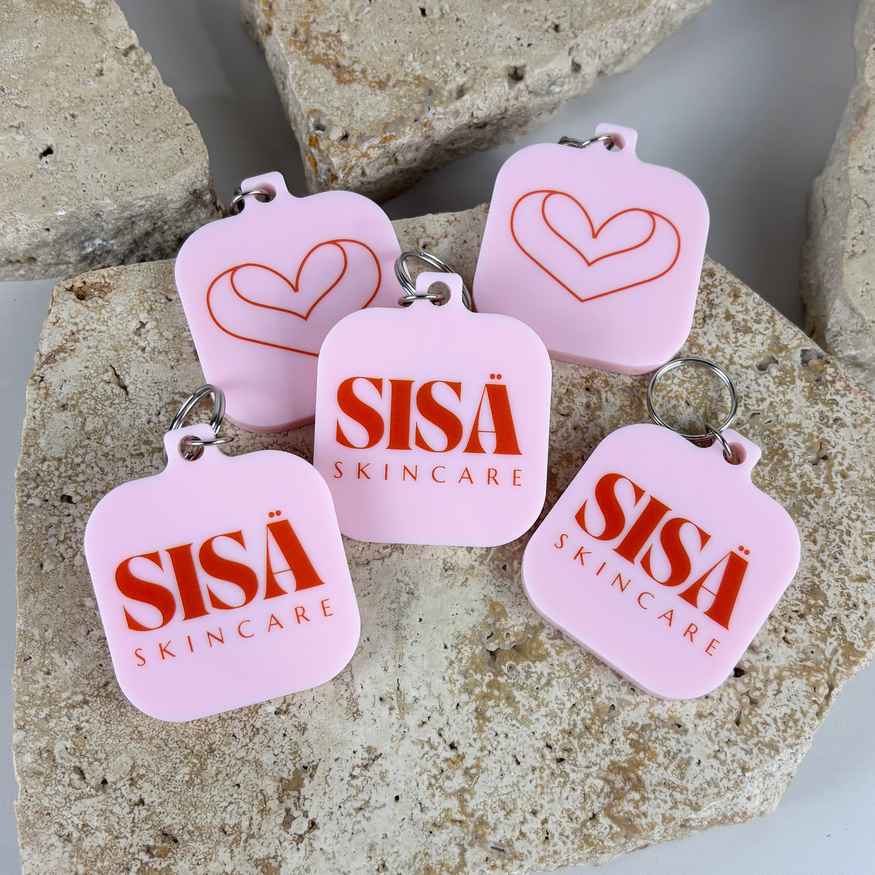 Pink acrylic keychains featuring the SISÄ Skincare logo and heart line-art designs in bold red, arranged on textured stone blocks for a clean, modern product display.