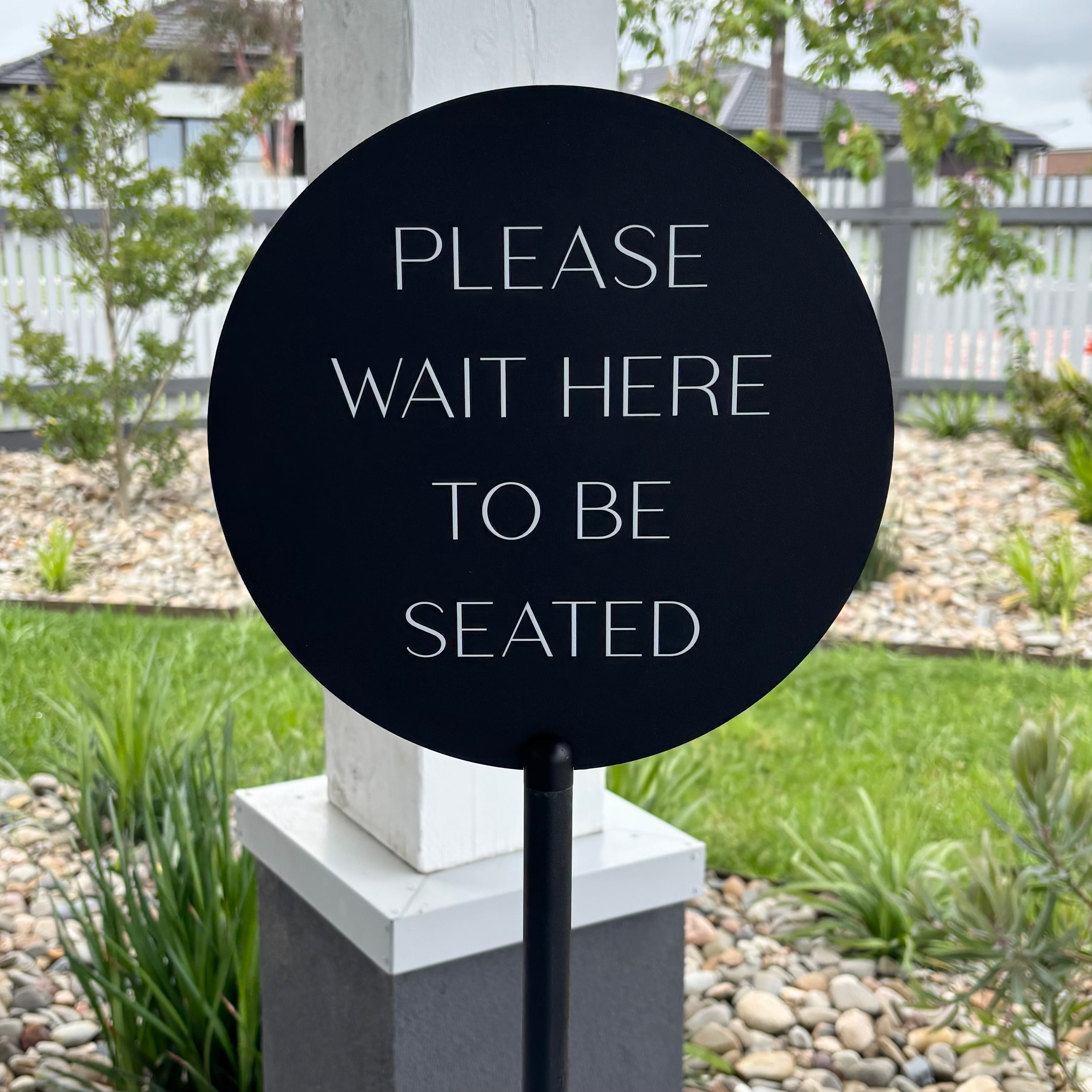 Round black acrylic hospitality sign on a pole reading ‘Please wait here to be seated’, custom made for restaurants and cafes, displayed outdoors near a garden.