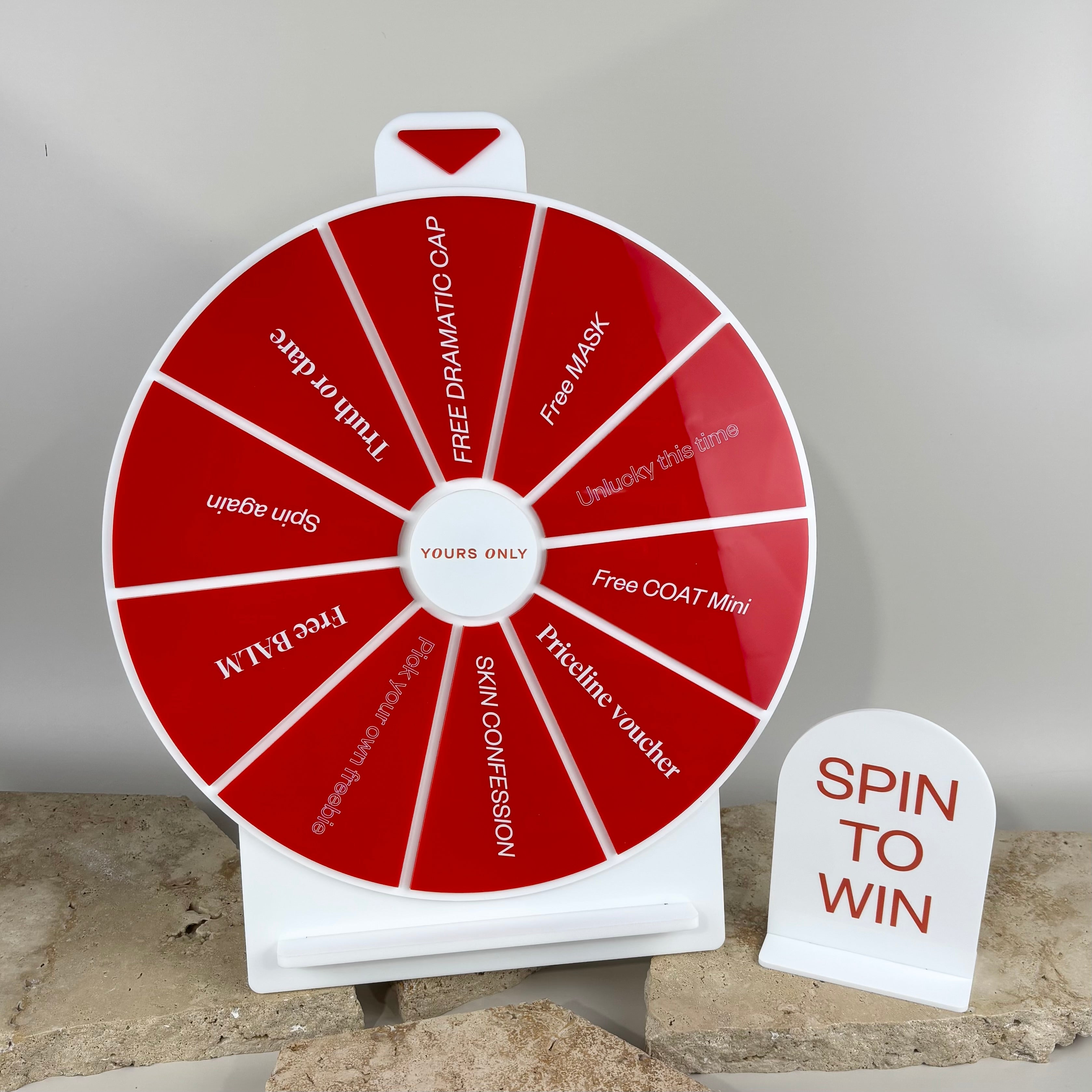 Custom red and white acrylic spin the wheel prize display with branded segments and a matching ‘Spin to Win’ counter sign, designed for retail promotions and salon giveaways, styled on natural stone tiles.