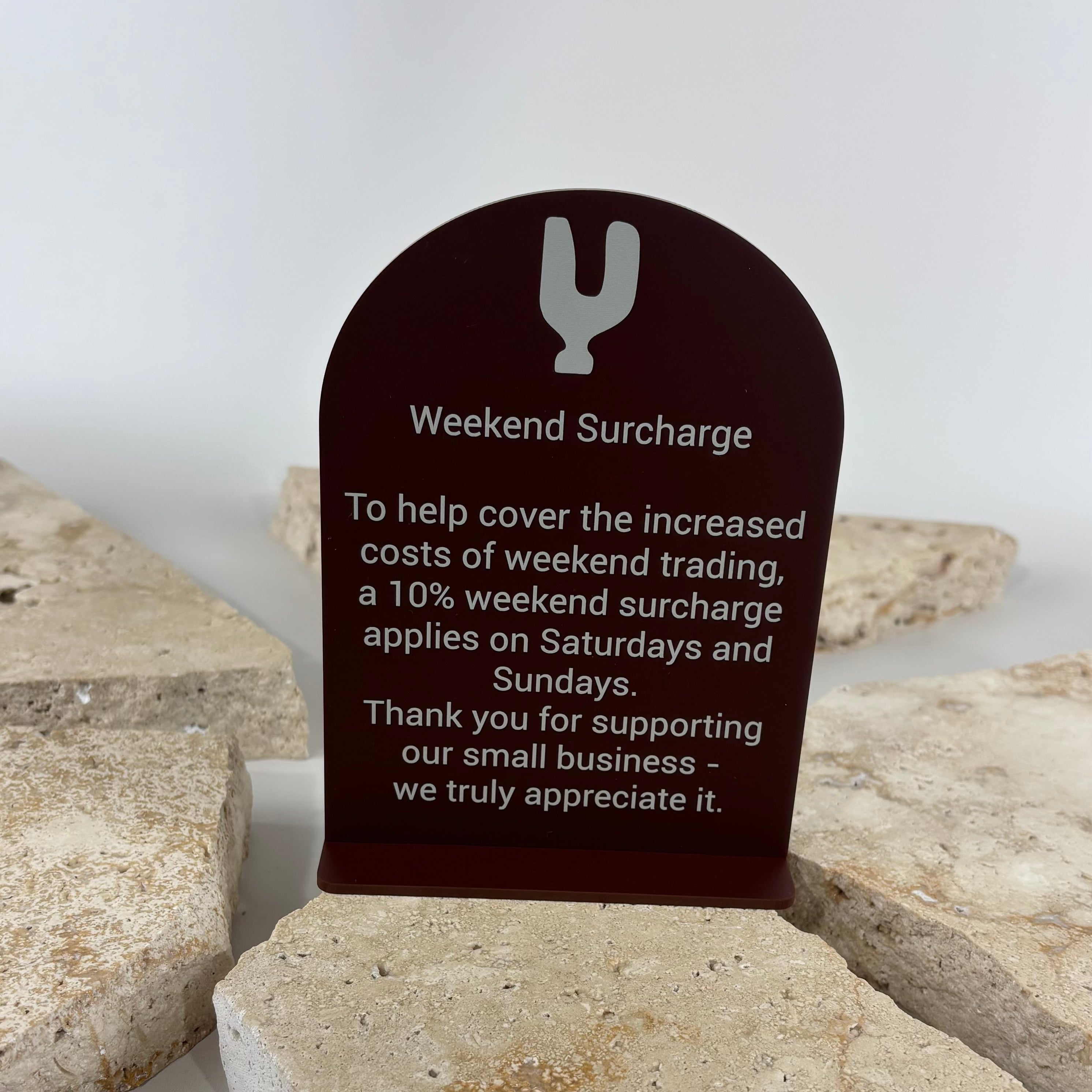 Custom acrylic weekend surcharge sign with UV printed white text on dark burgundy acrylic. Freestanding countertop business sign explaining a 10% weekend surcharge for Saturdays and Sundays, ideal for cafés, restaurants, bakeries, retail stores and hospitality businesses.