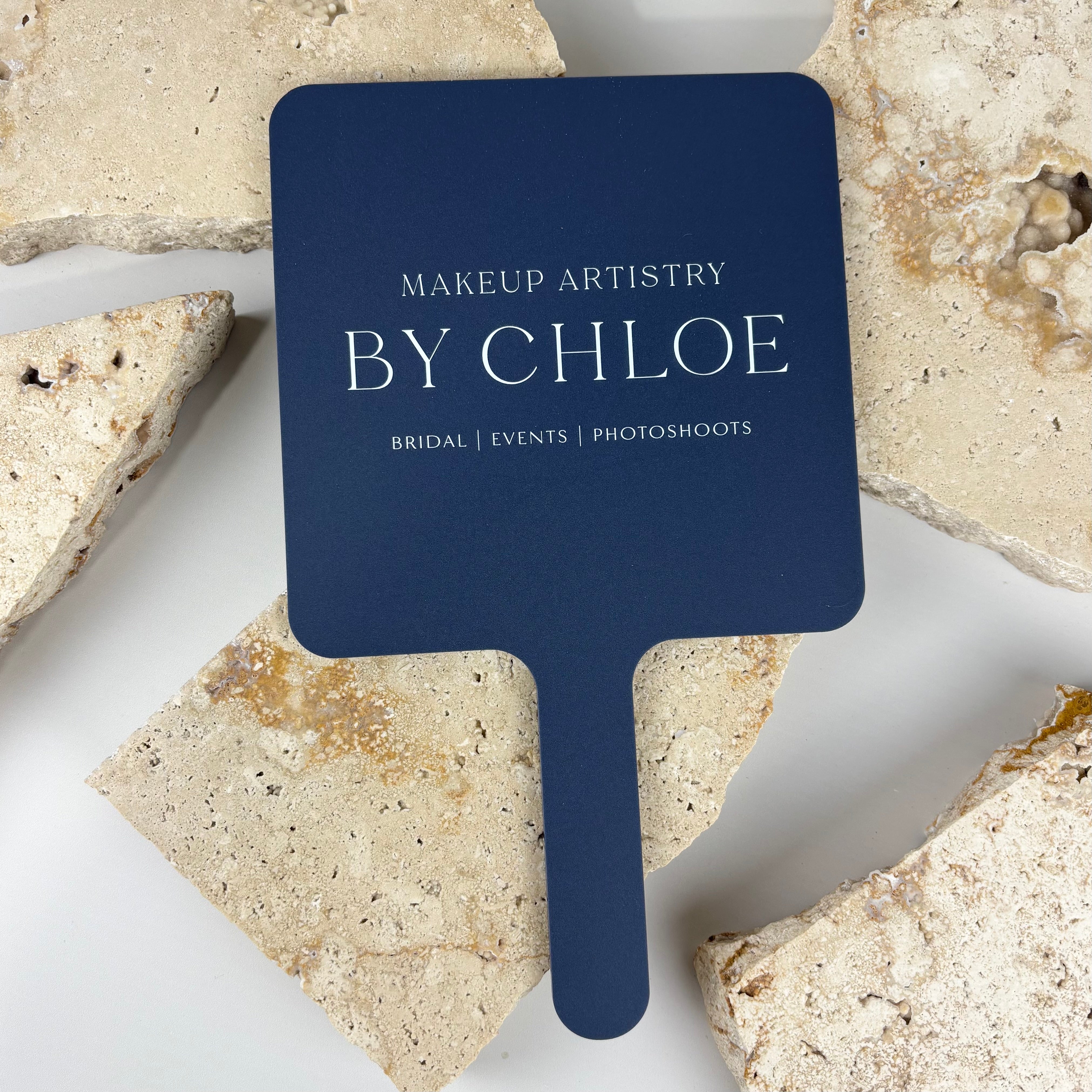 Navy blue acrylic handheld mirror with square rounded shape, featuring white logo text reading Makeup Artistry by Chloe, styled on a neutral stone background