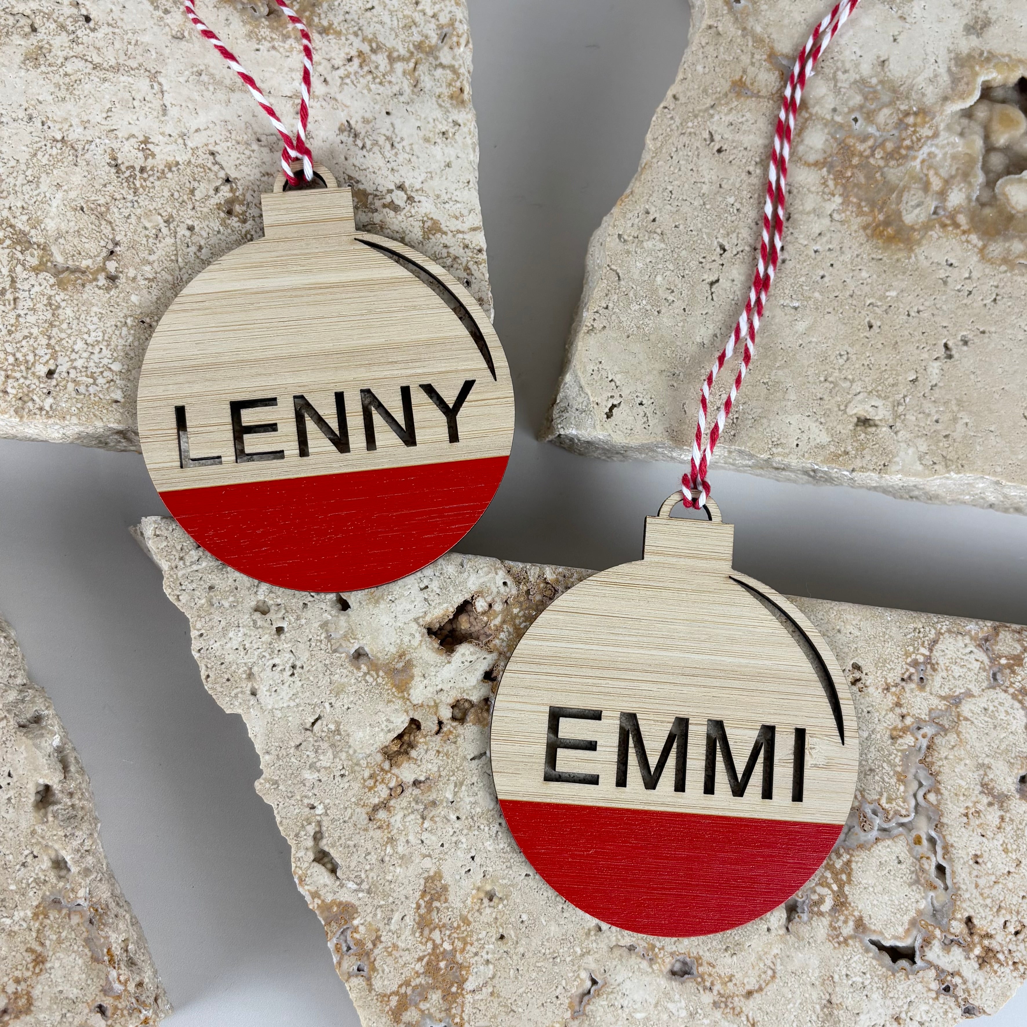 personalised wooden christmas bauble ornaments with red painted base and engraved names lenny and emmi on stone background, custom laser cut timber holiday decorations australia made eco friendly keepsake