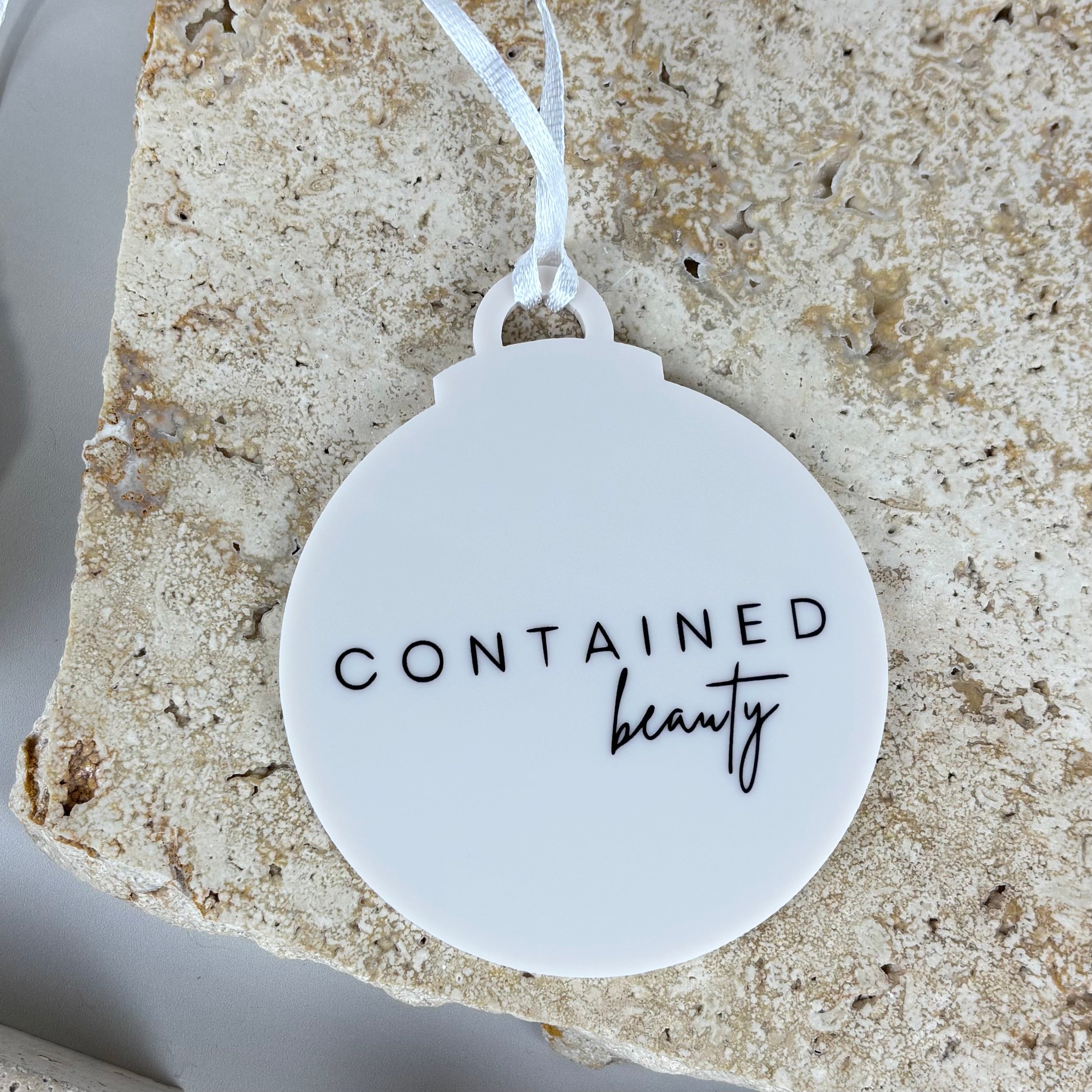 White acrylic Christmas bauble with engraved black logo text “Contained Beauty”, minimalist round ornament with white ribbon, custom branded business Christmas decoration.