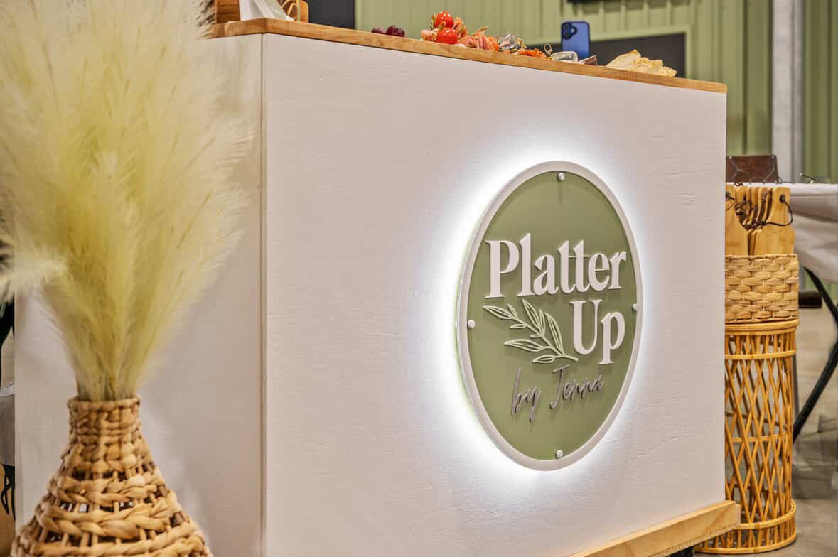 Custom round backlit business sign installed on a white catering counter for Platter Up by Jenna, featuring 3D acrylic lettering on a sage green backing, laser cut and illuminated for event display.