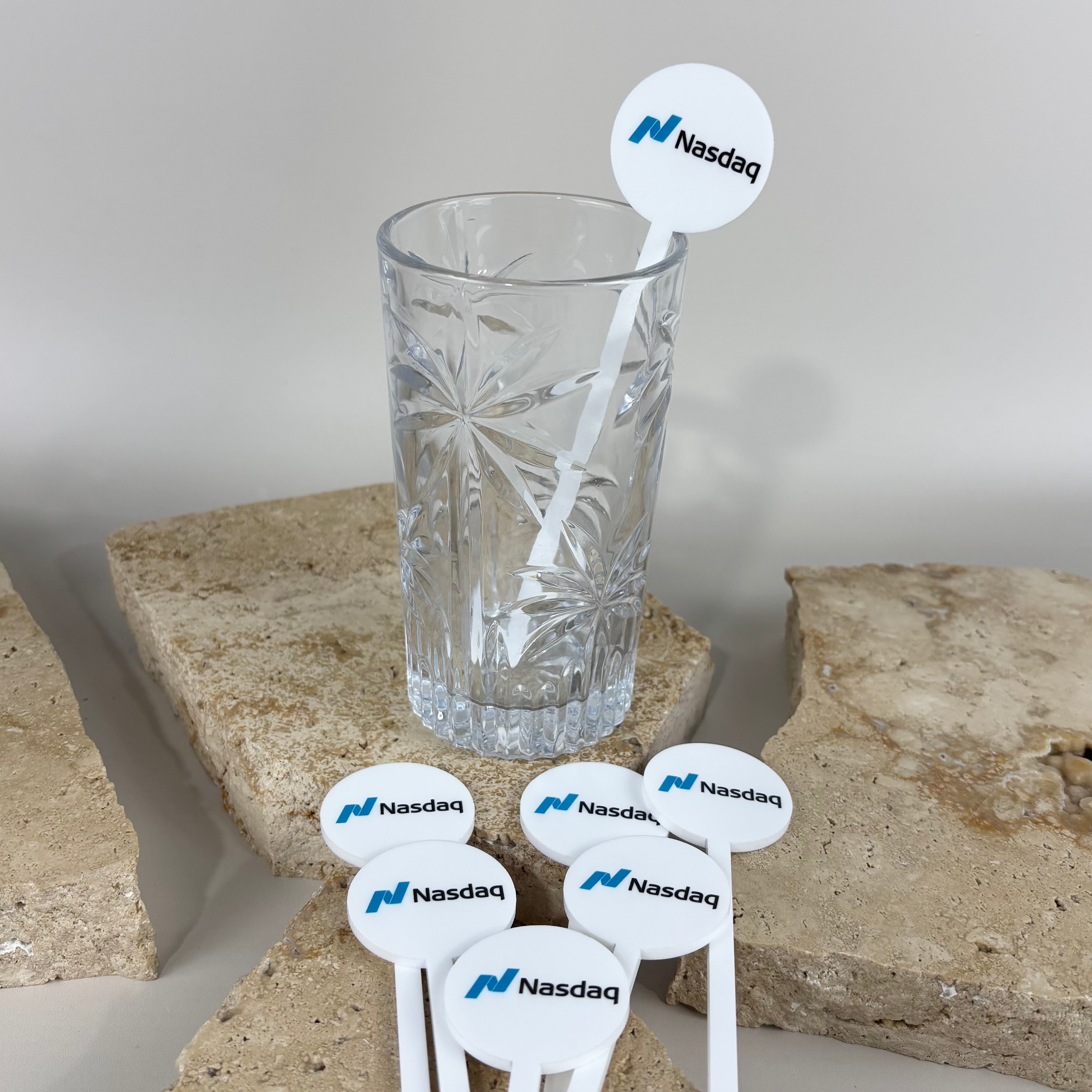 Custom white acrylic drink stirrers with blue Nasdaq logo, shown inside a crystal glass and displayed on stone backdrop, branded cocktail accessories for corporate events and promotional gifting.