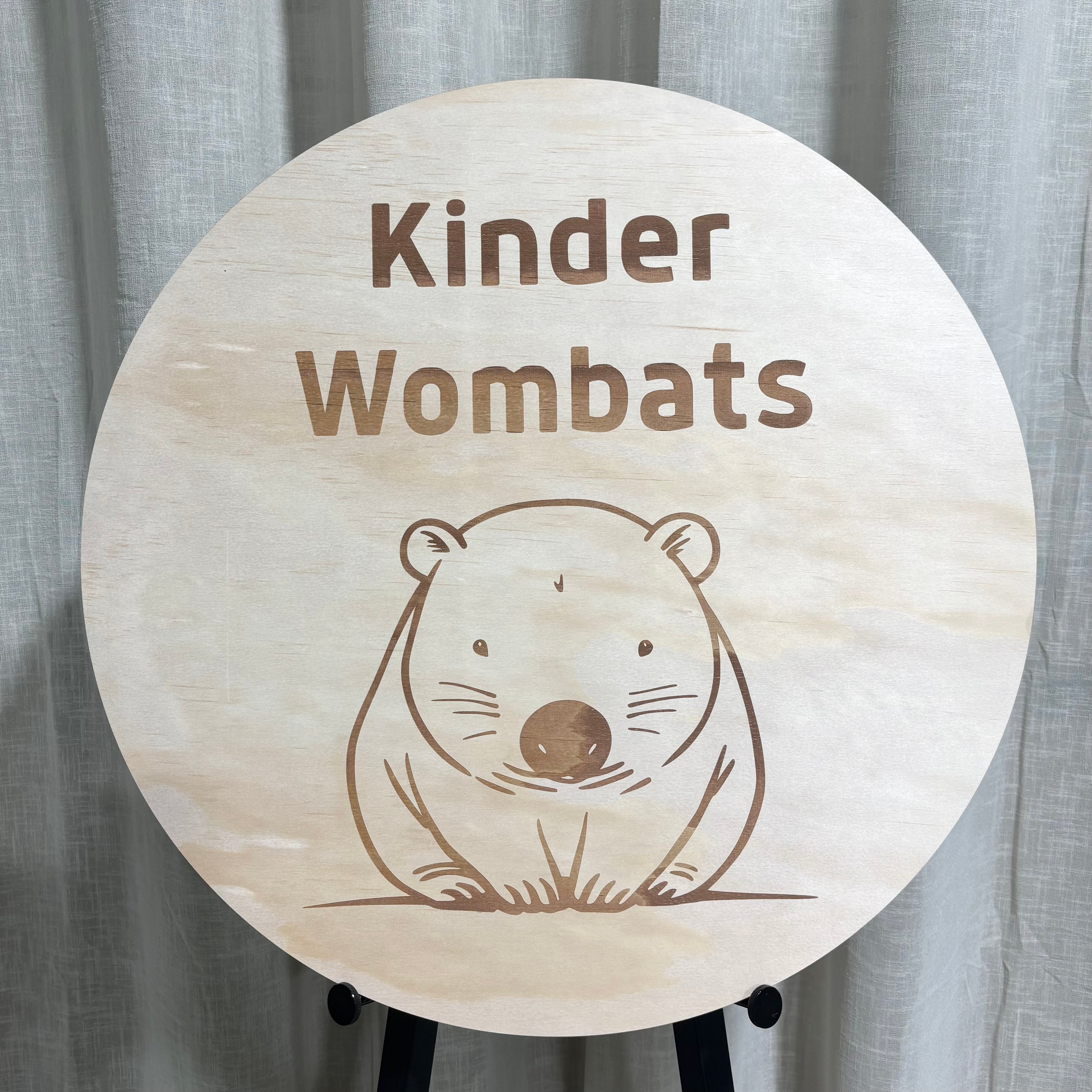 Round wooden kindergarten sign engraved with ‘Kinder Wombats’ text and a cute wombat illustration, natural timber finish displayed on a stand indoors.