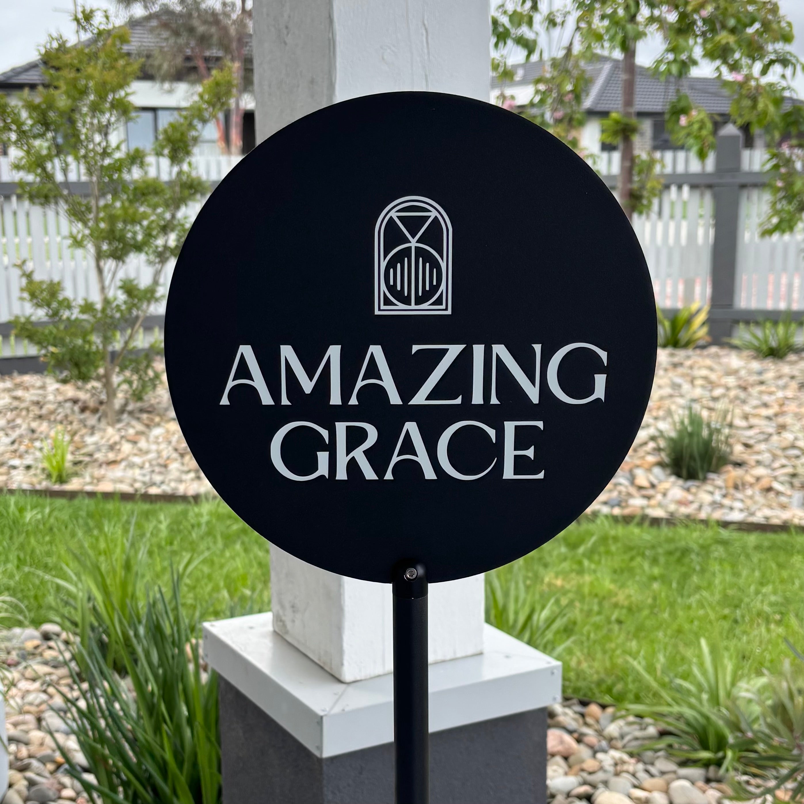 Custom round black acrylic business sign on a pole featuring the Amazing Grace logo and text, designed for cafes and venues, photographed outdoors near a landscaped garden.