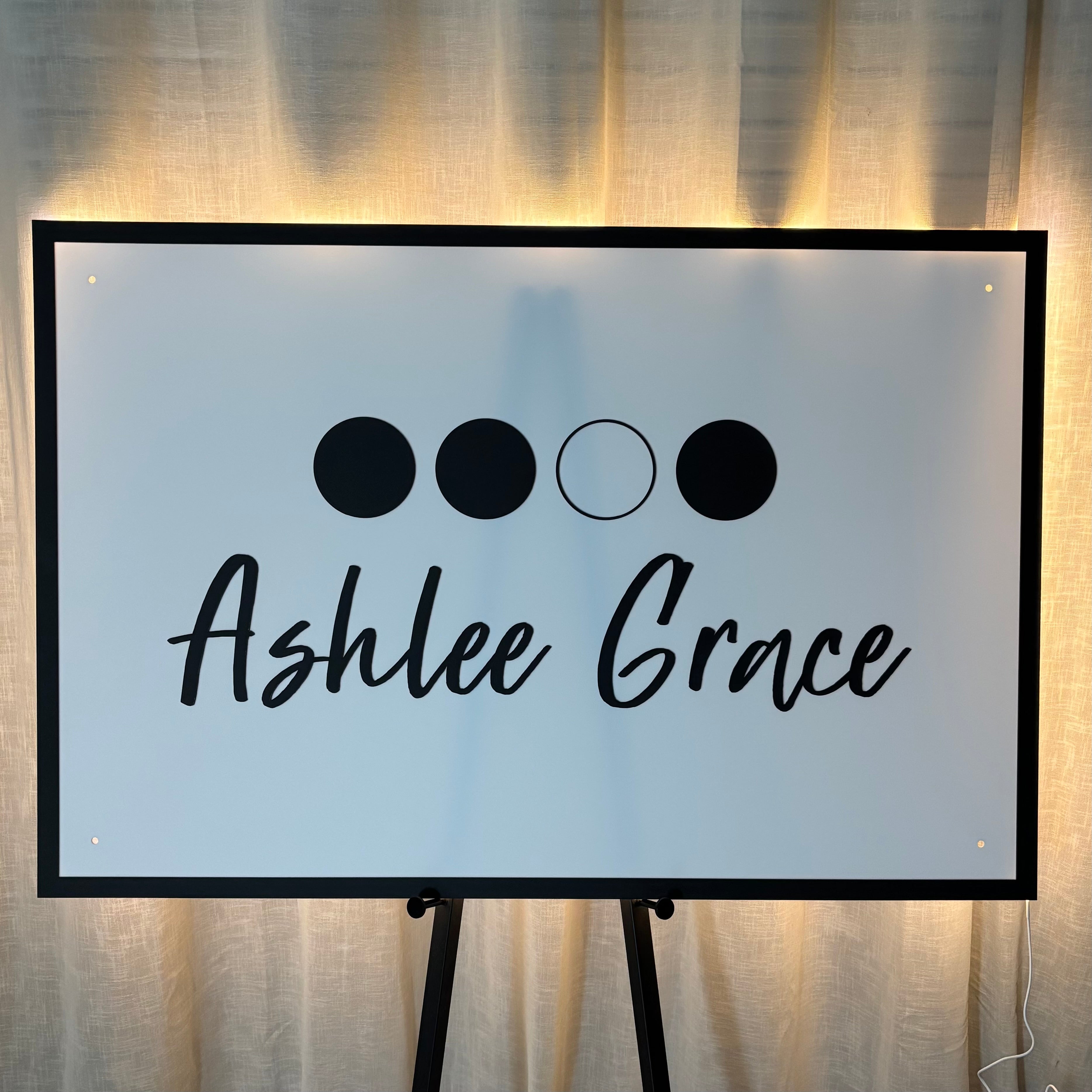 Rectangular backlit acrylic business sign on a black metal floor stand featuring the name “Ashlee Grace” in black script, illuminated with warm LED lighting and positioned in front of neutral curtains for a modern beauty salon or clinic signage display.
