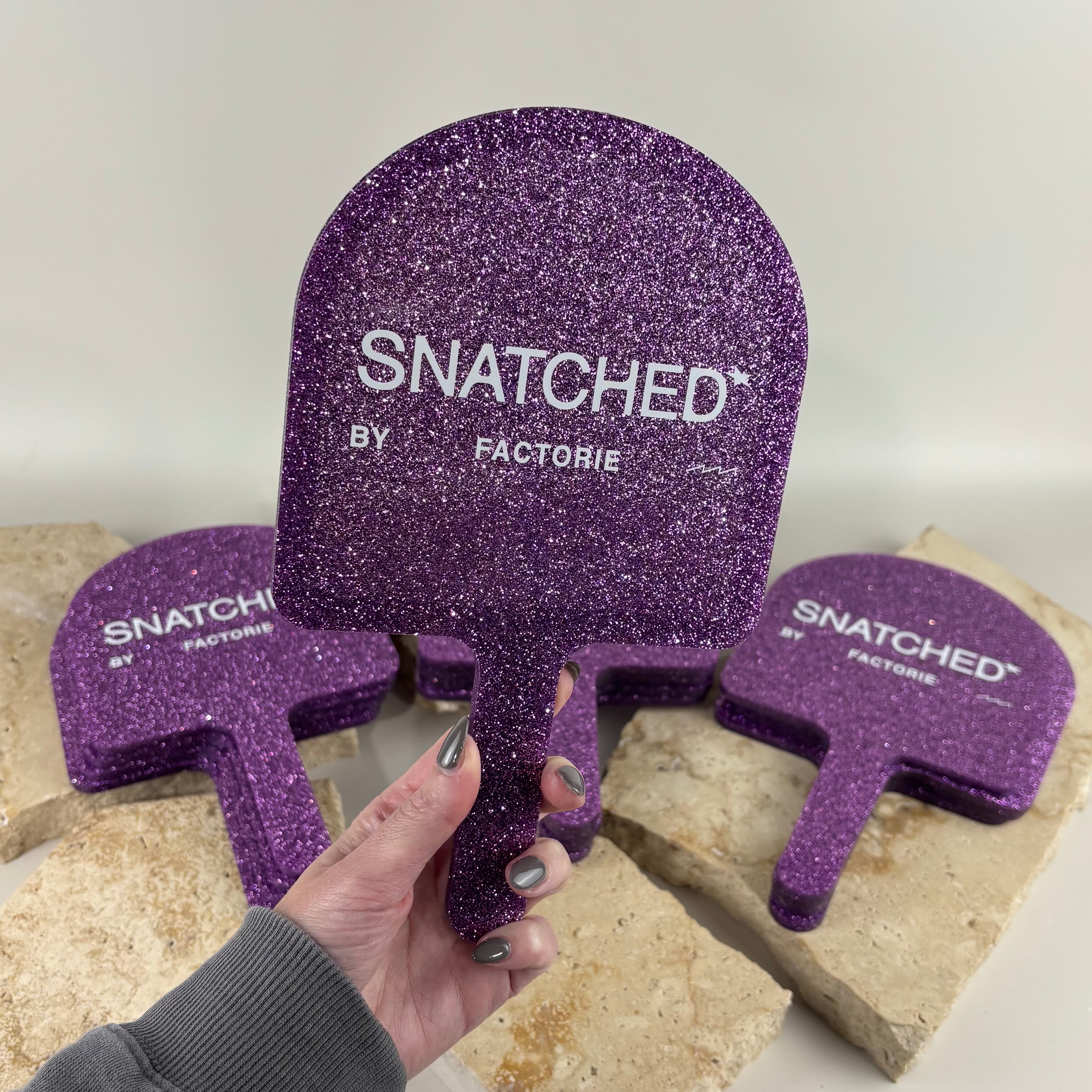 Custom purple glitter handheld salon mirror with UV printed white branding for Snatched by Factorie. Premium glitter acrylic hand mirror for beauty salons, cosmetic clinics, makeup artists and lash studios, perfect for client treatments, selfies, retail displays and branded salon accessories.
