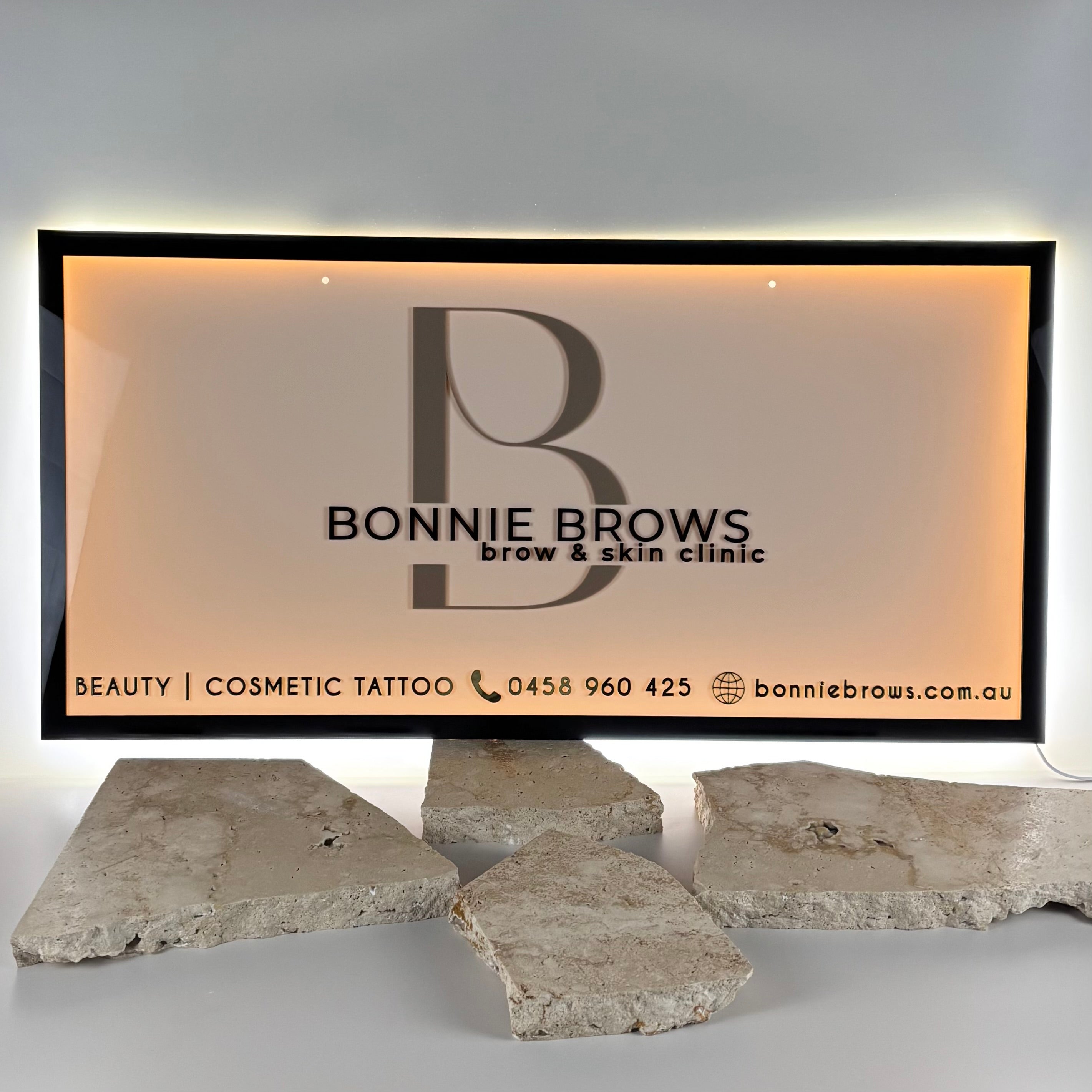 Illuminated acrylic business sign for “Bonnie Brows Brow & Skin Clinic” featuring a large B logo, contact details, and website bonniebrows.com.au, displayed with a soft backlight and placed on textured stone slabs.