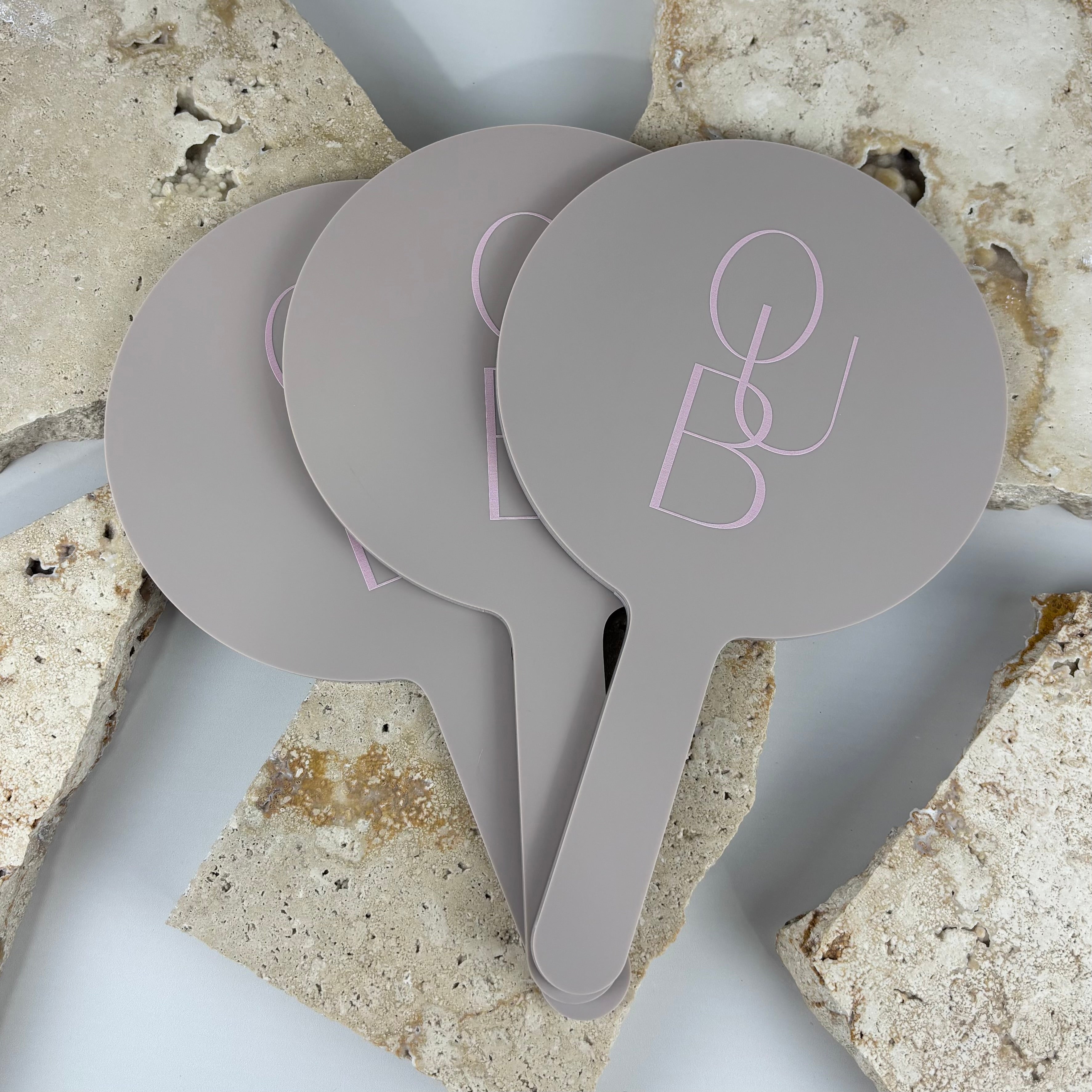 Neutral taupe acrylic handheld mirrors stacked together, featuring a minimalist pink B&J logo, styled on a textured stone backdrop
