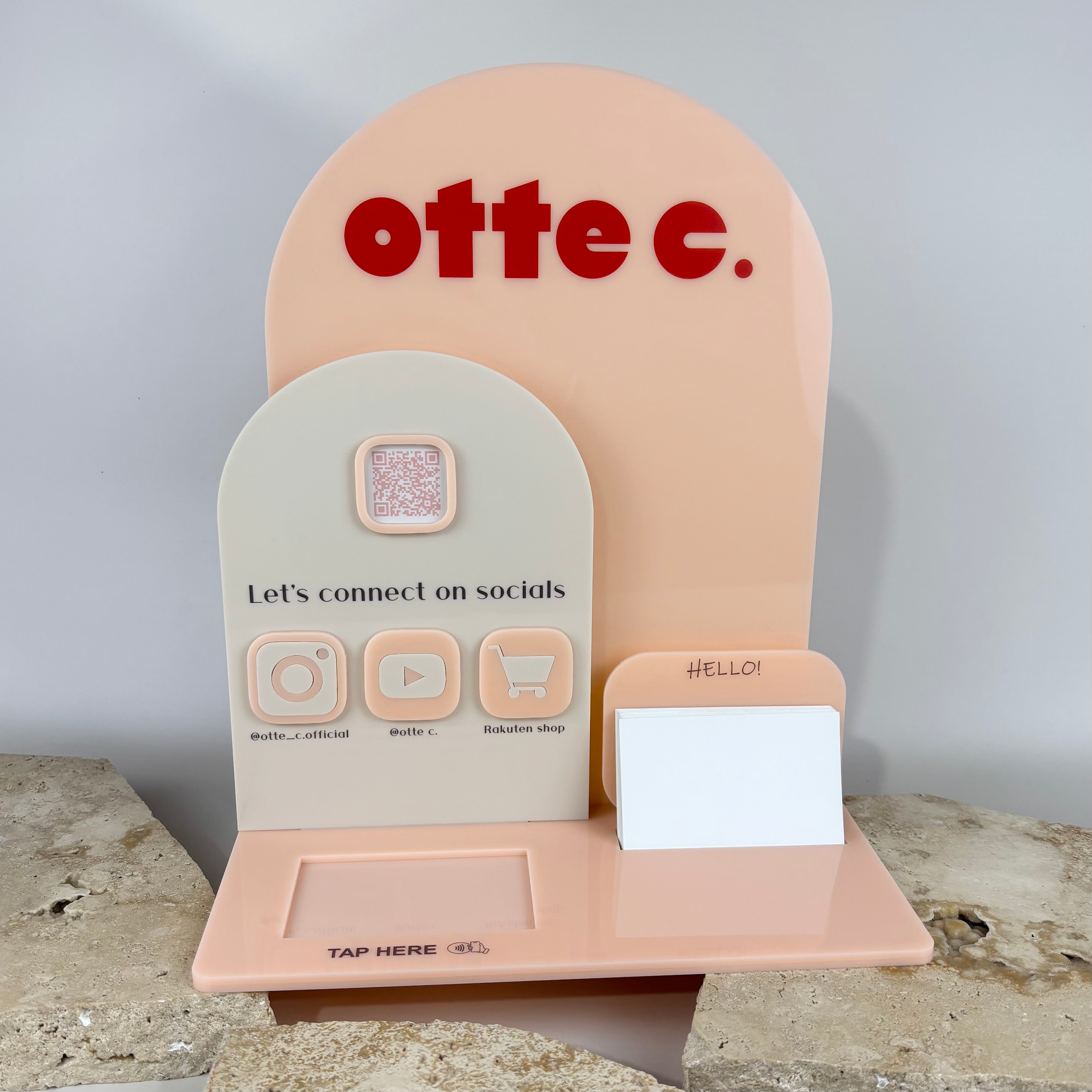Custom peach and cream acrylic NFC business station for “otte c.” featuring layered arch design, QR code, social media icons for Instagram, YouTube, and Rakuten shop, a “Tap Here” NFC area, and a built-in business card holder, displayed on stone slabs.