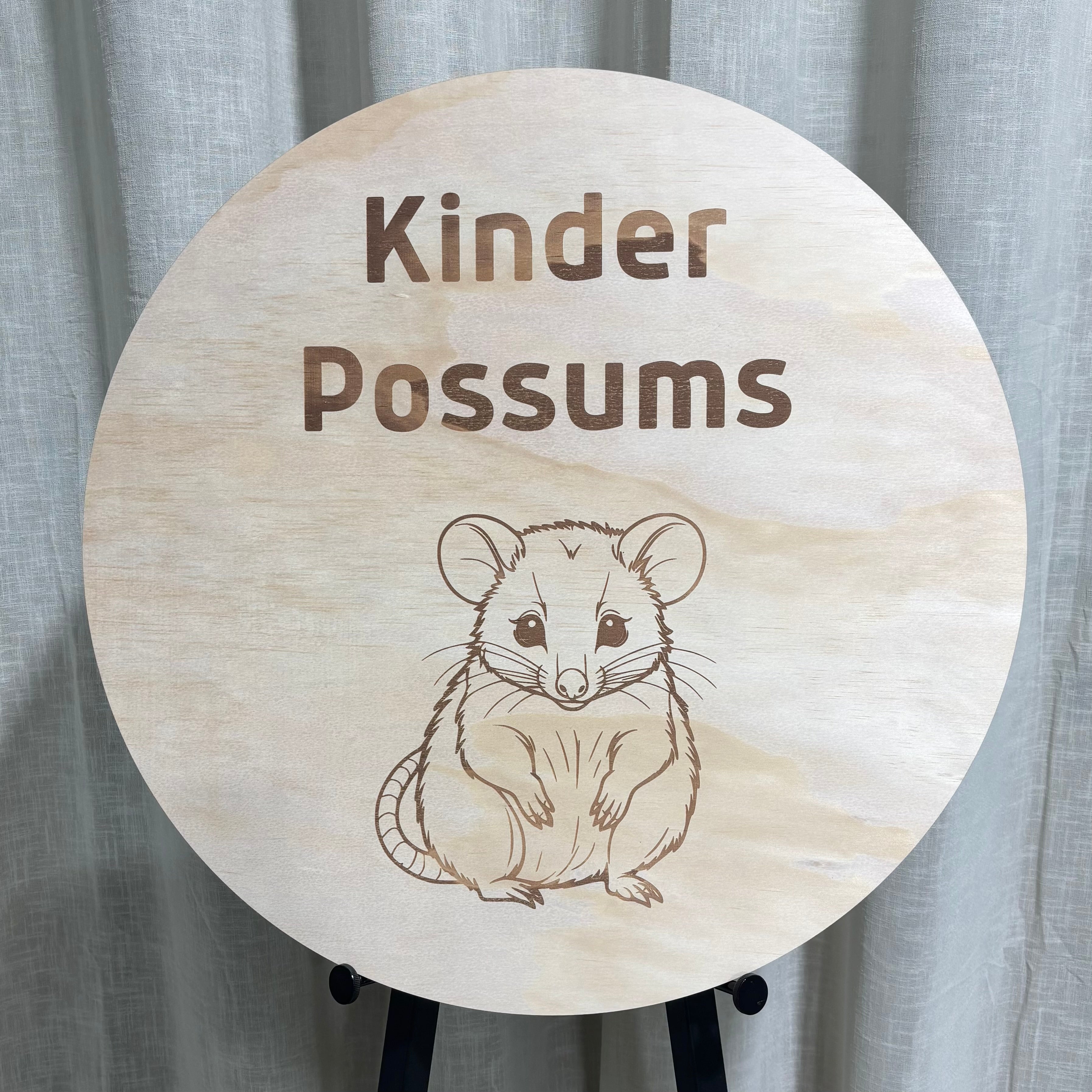Round wooden kindergarten sign with engraved ‘Kinder Possums’ text and illustrated possum design, displayed on an easel against a neutral curtain background.
