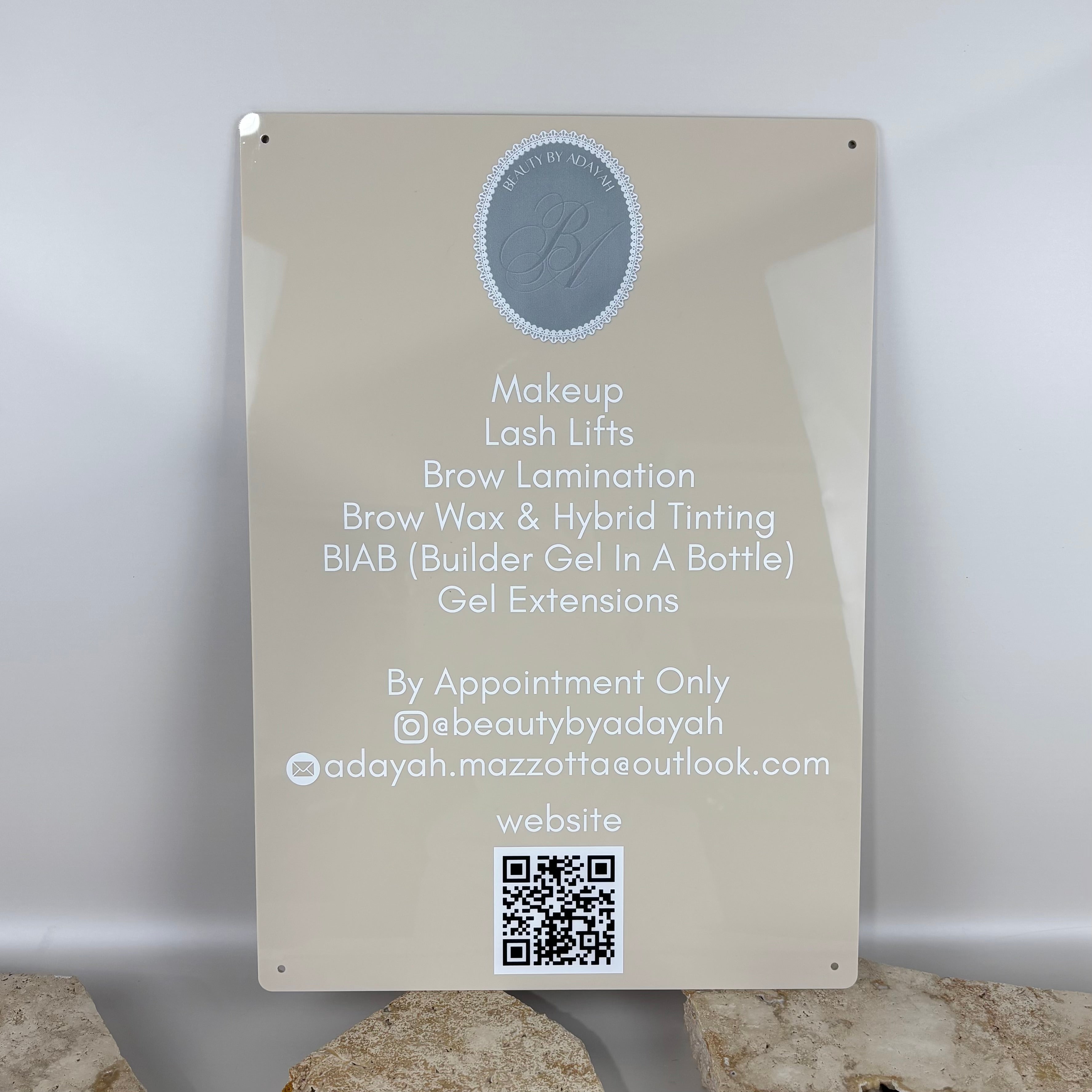 Wall-mounted acrylic beauty services sign in neutral beige with white text, listing makeup, lash lifts, brow lamination, BIAB and gel extensions, featuring QR code, Instagram handle and contact details for a beauty salon.