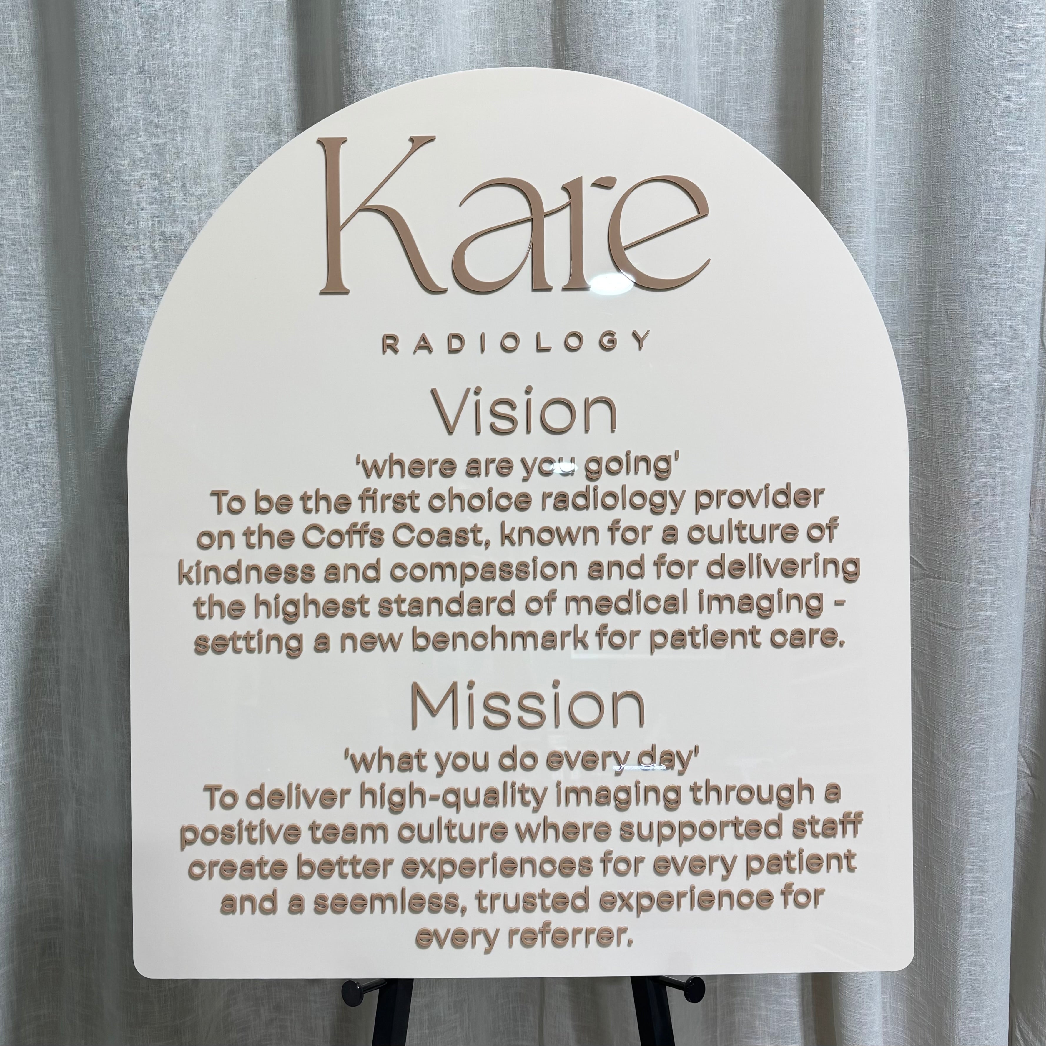 Custom arched acrylic business sign for Kaie Radiology featuring layered 3D acrylic lettering in a neutral beige and white colour palette. Modern radiology clinic reception sign with vision and mission statement text, premium UV printed and raised acrylic details, ideal for medical centres, healthcare clinics, reception areas and professional office branding in Australia.