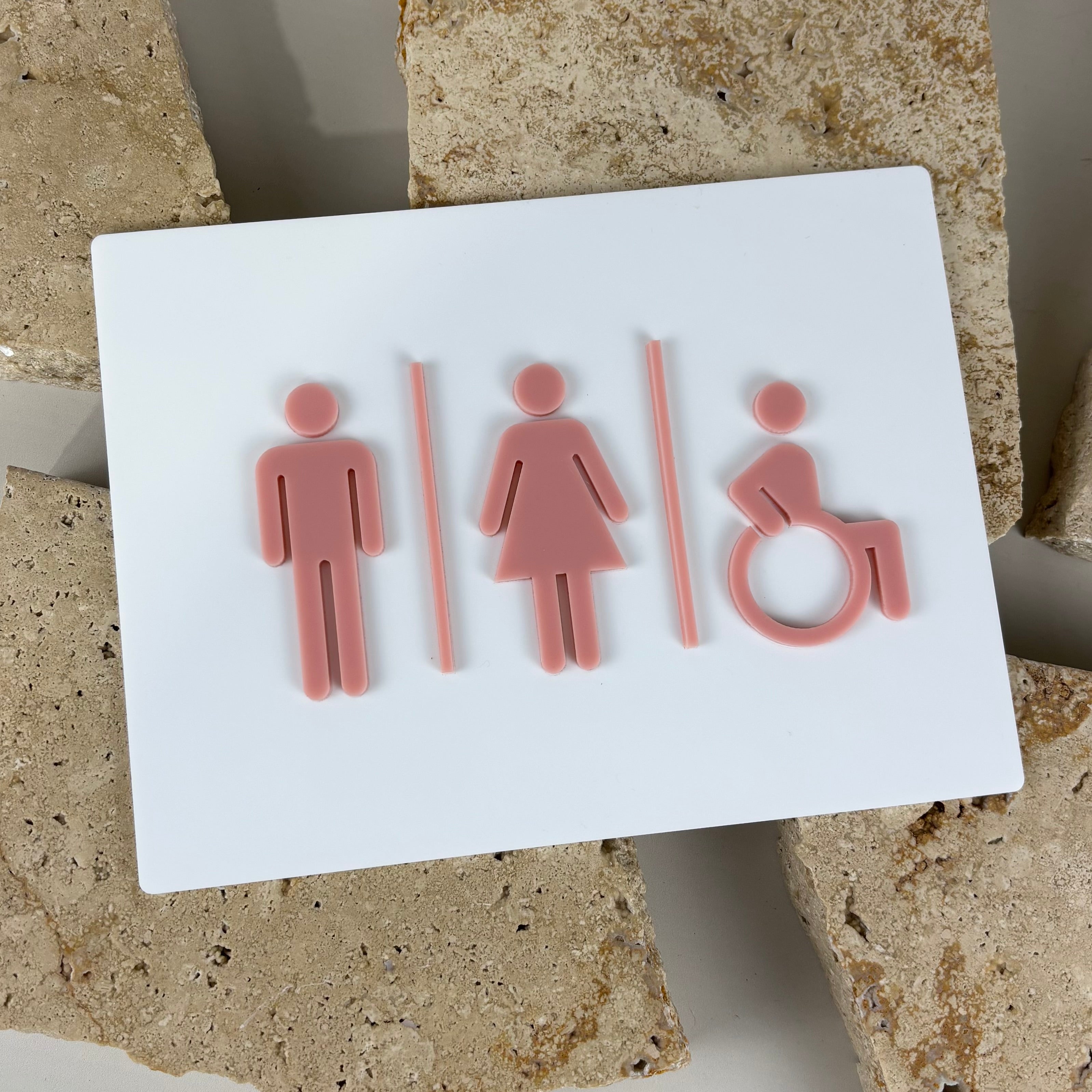 Modern acrylic bathroom sign with male, female and wheelchair symbols in blush pink on white background, commercial restroom signage
