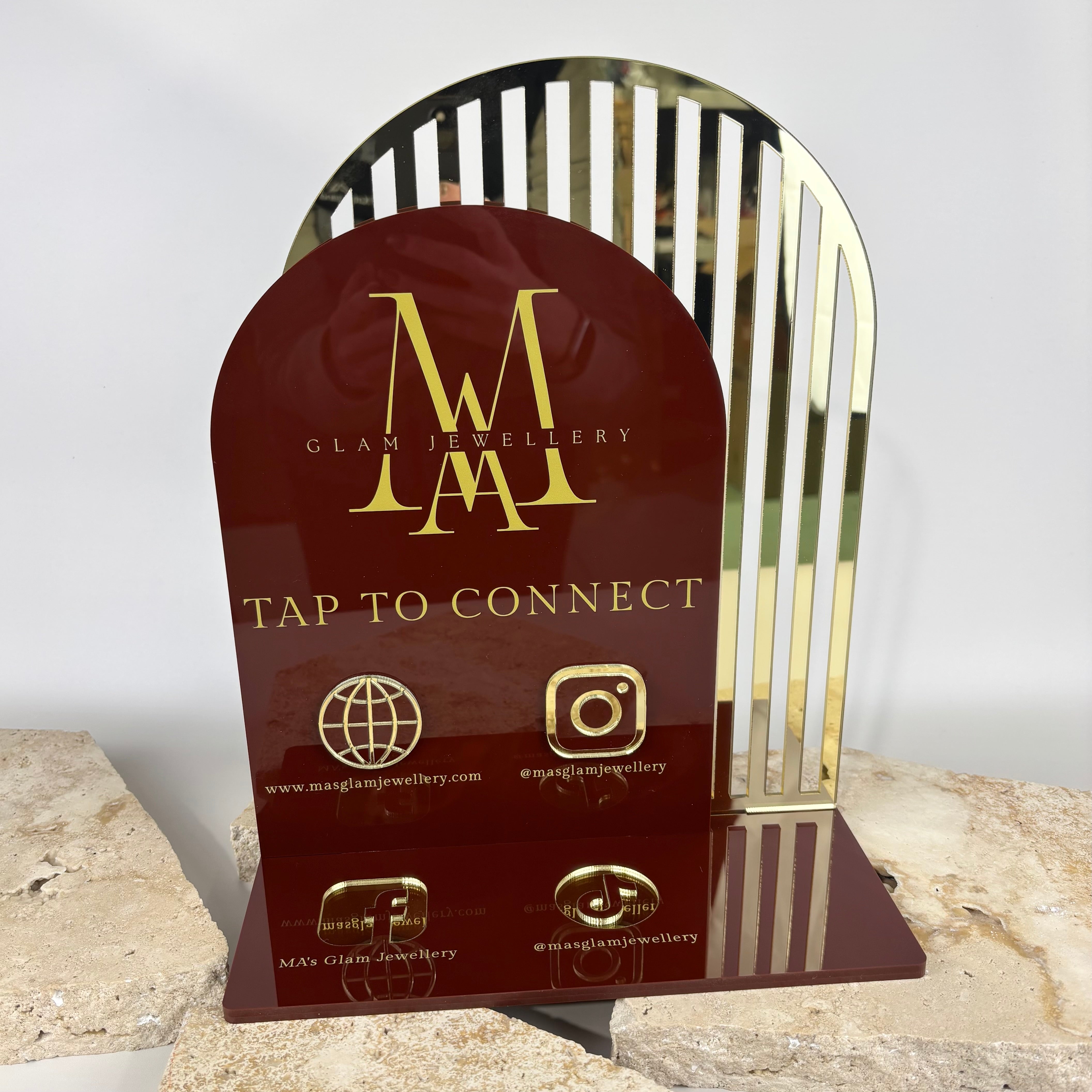Luxury burgundy and gold acrylic NFC business display for MA’s Glam Jewellery, featuring “Tap to Connect” text, website and social media icons for Instagram, Facebook, and TikTok, with a gold striped mirror backing, displayed on stone slabs.
