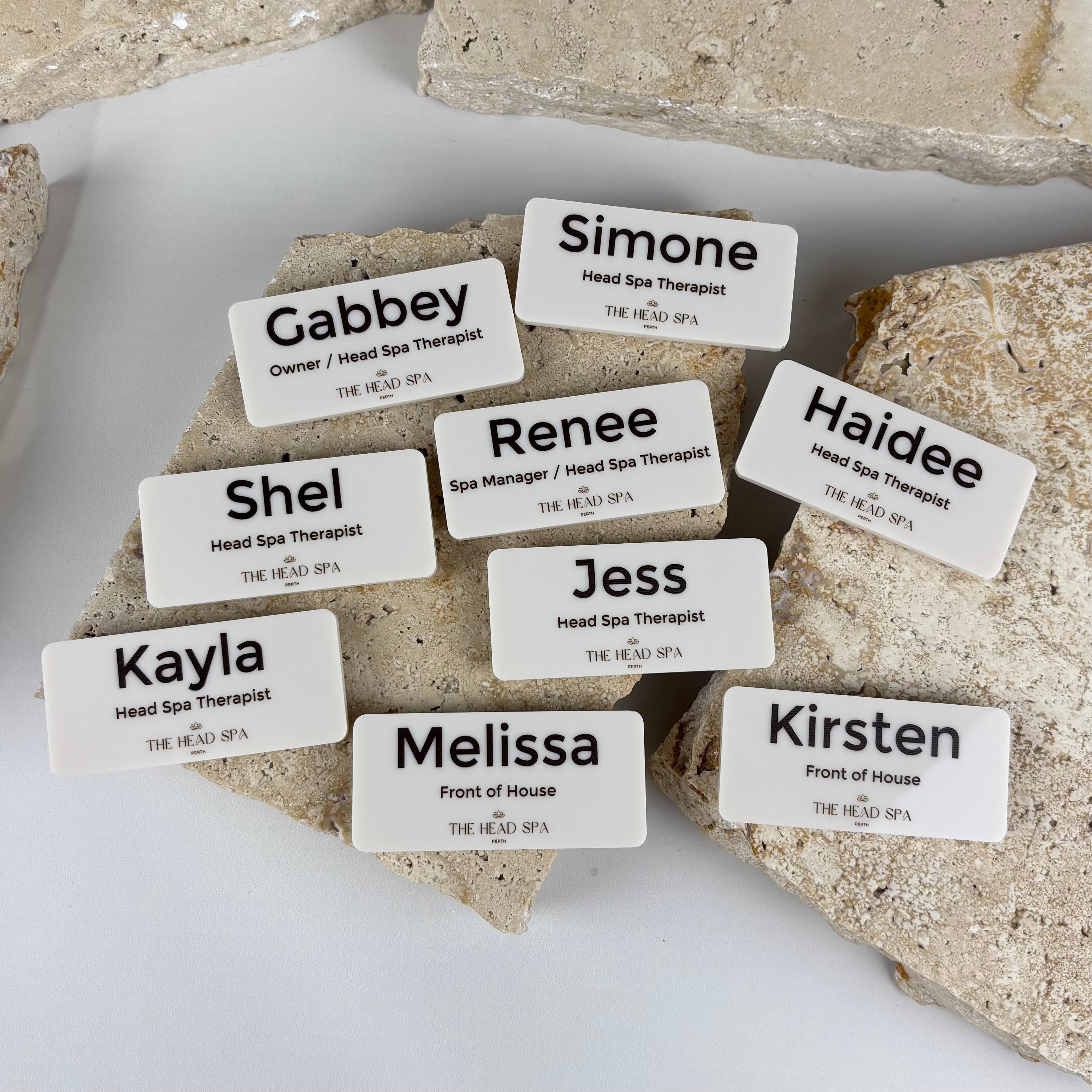 Custom white acrylic name badges with black engraved names and job titles for spa staff, including head spa therapists and front of house, styled on a neutral stone background