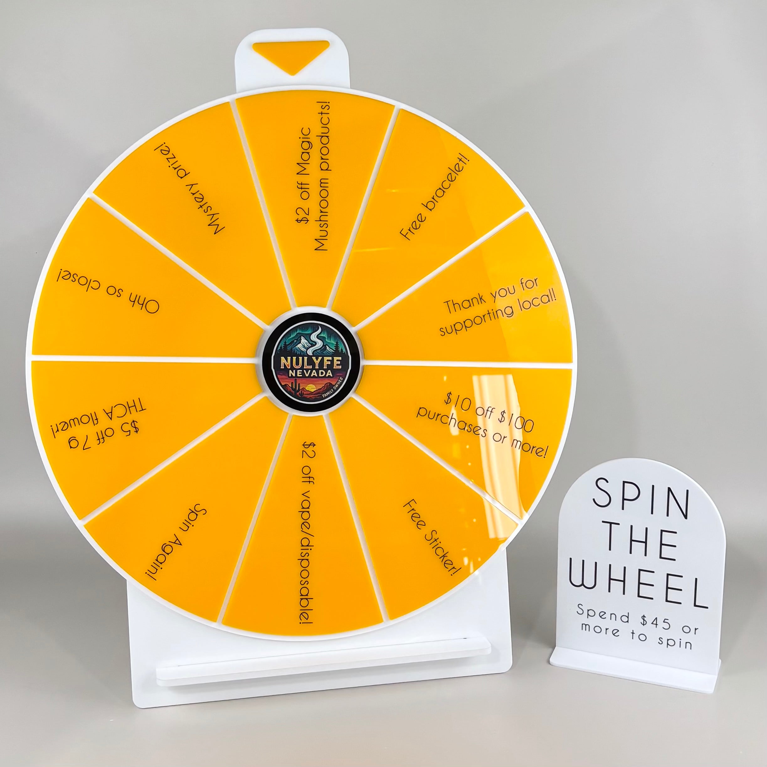 Custom yellow acrylic spin the wheel prize display for retail stores, featuring branded Nulyfe Nevada centre logo and giveaway sections for discounts, free products and rewards, paired with white acrylic “Spin the Wheel – Spend $45 or more to spin” sign on grey background.