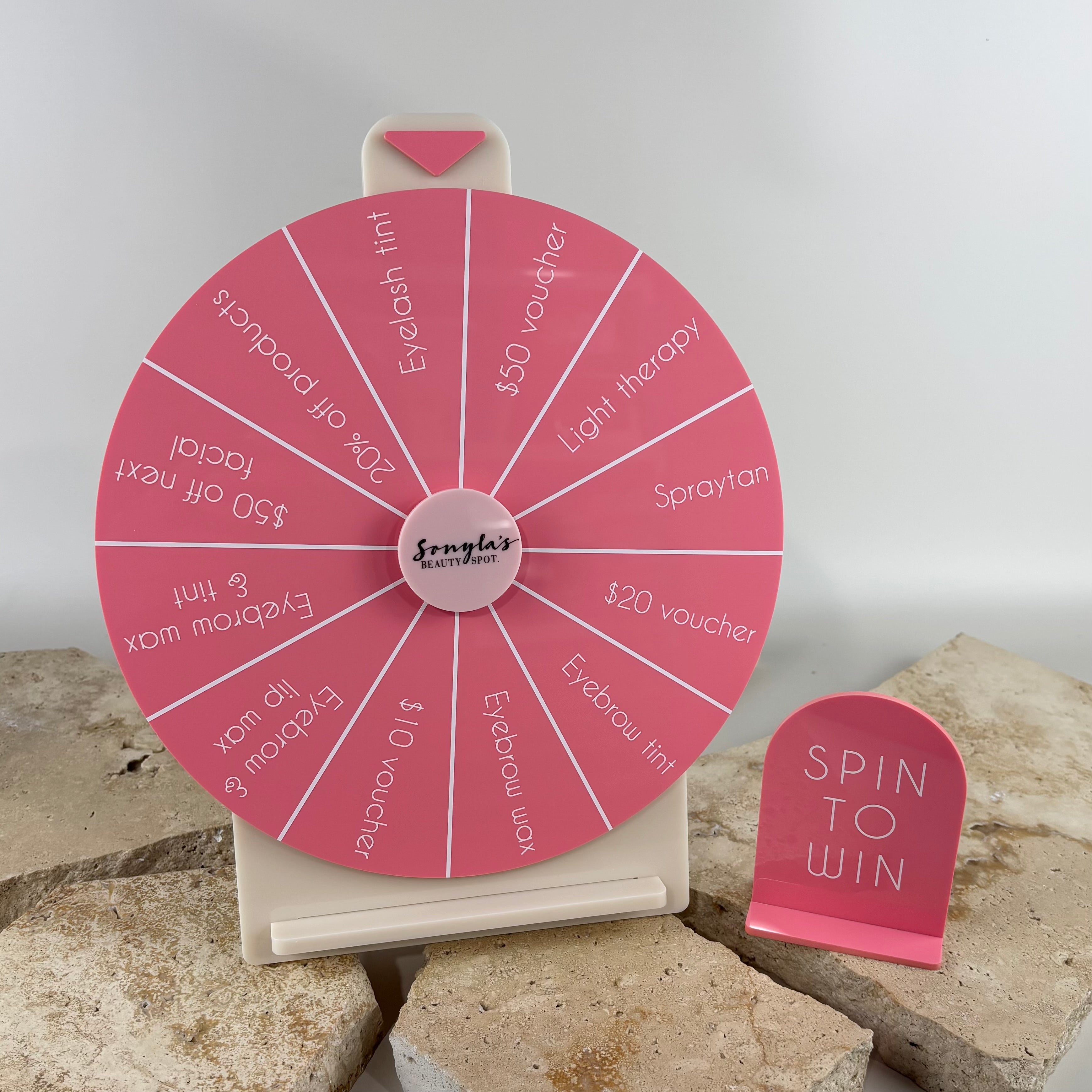 Custom acrylic spin to win prize wheel for salons and retail stores featuring branded pink spinning wheel with beauty treatment prizes, vouchers, eyebrow wax, eyelash tint and spray tan rewards. Promotional prize wheel display with matching spin to win sign for customer engagement, giveaways and in-store marketing.
