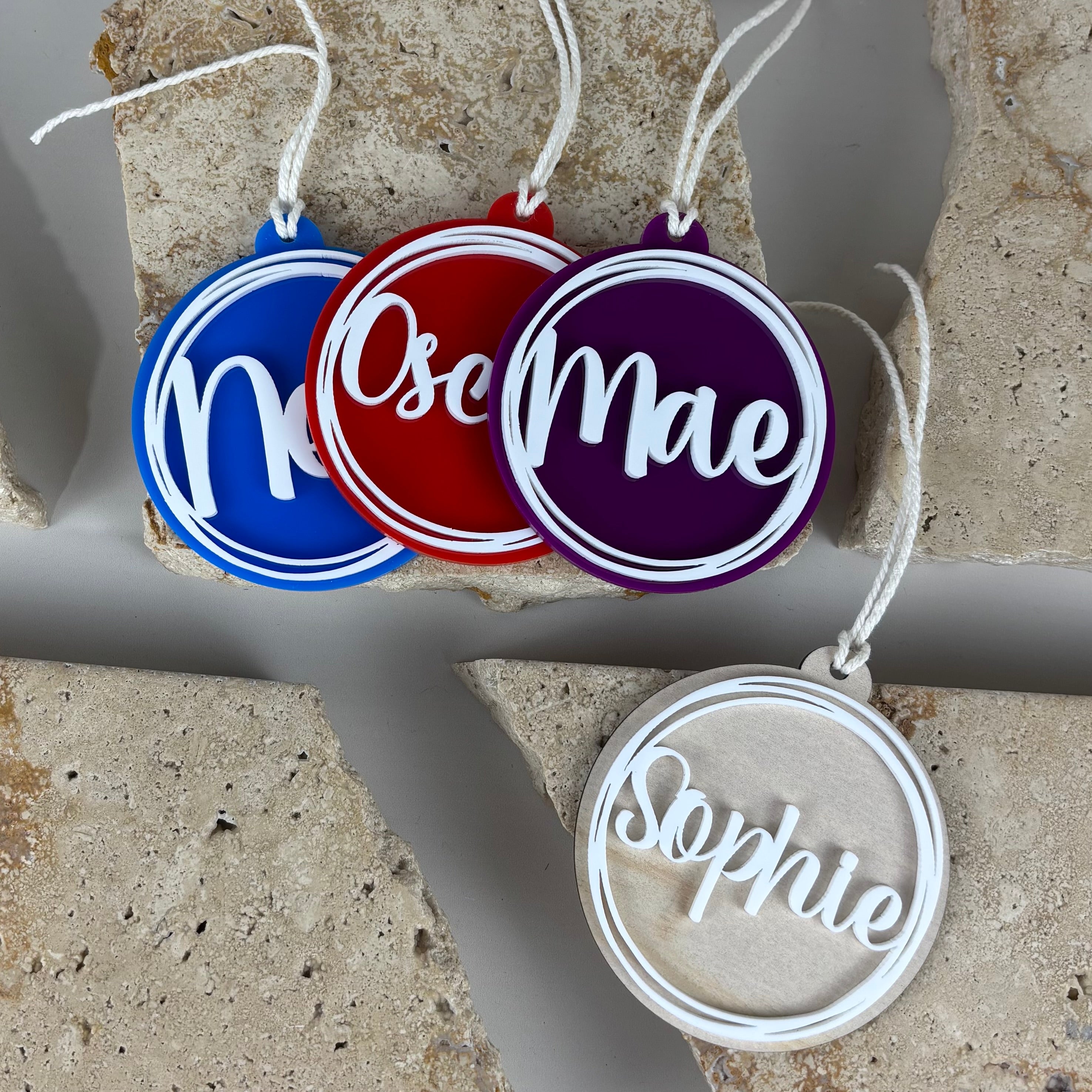 Personalised acrylic Christmas baubles in blue, red, purple and natural wood, layered round ornaments with raised white script names “Noah”, “Oscar”, “Mae” and “Sophie”.