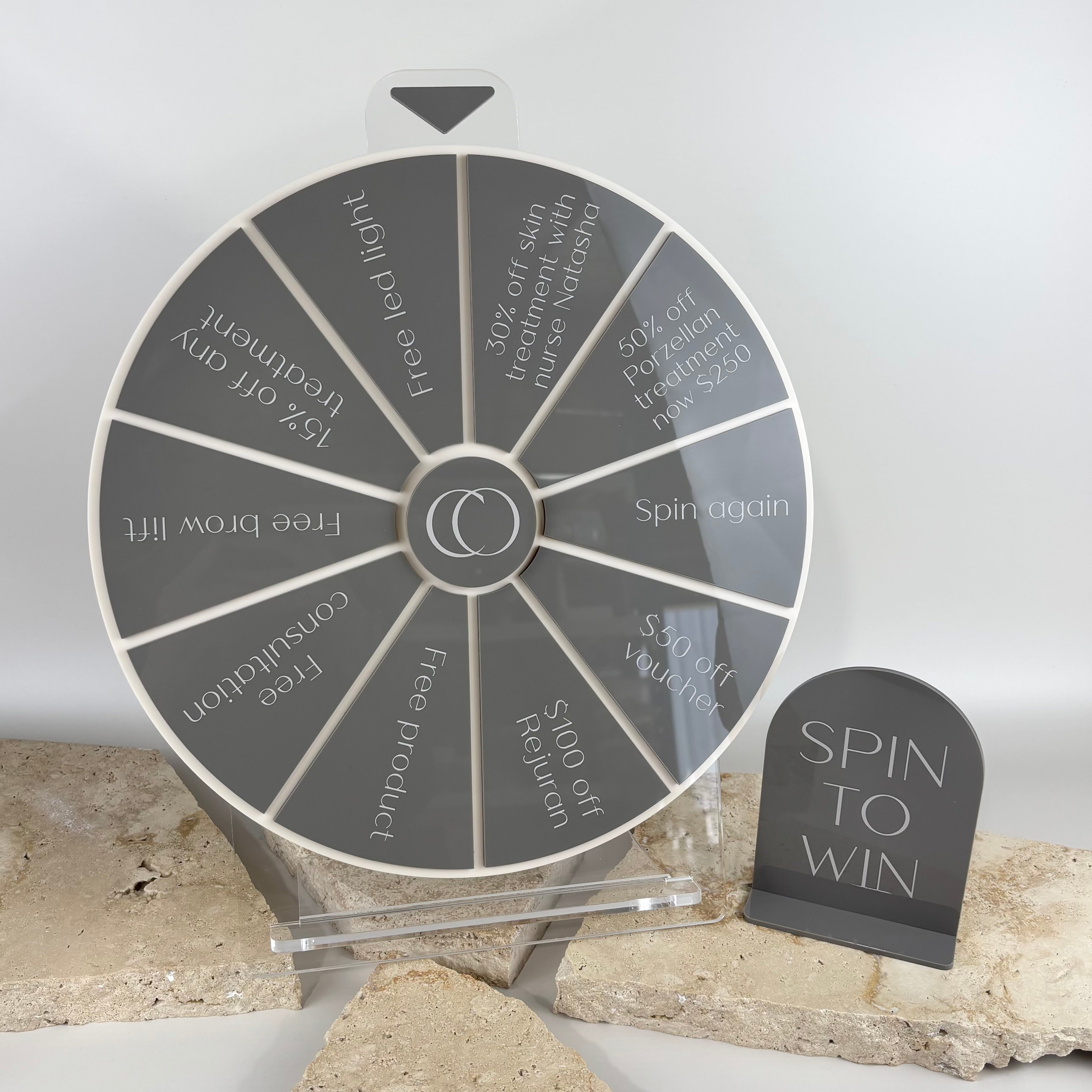 Custom grey acrylic spin to win prize wheel for clinics and beauty salons, featuring engraved giveaway sections like discounts, free treatments, vouchers and product prizes, paired with matching grey “Spin to Win” counter sign, displayed on stone backdrop.