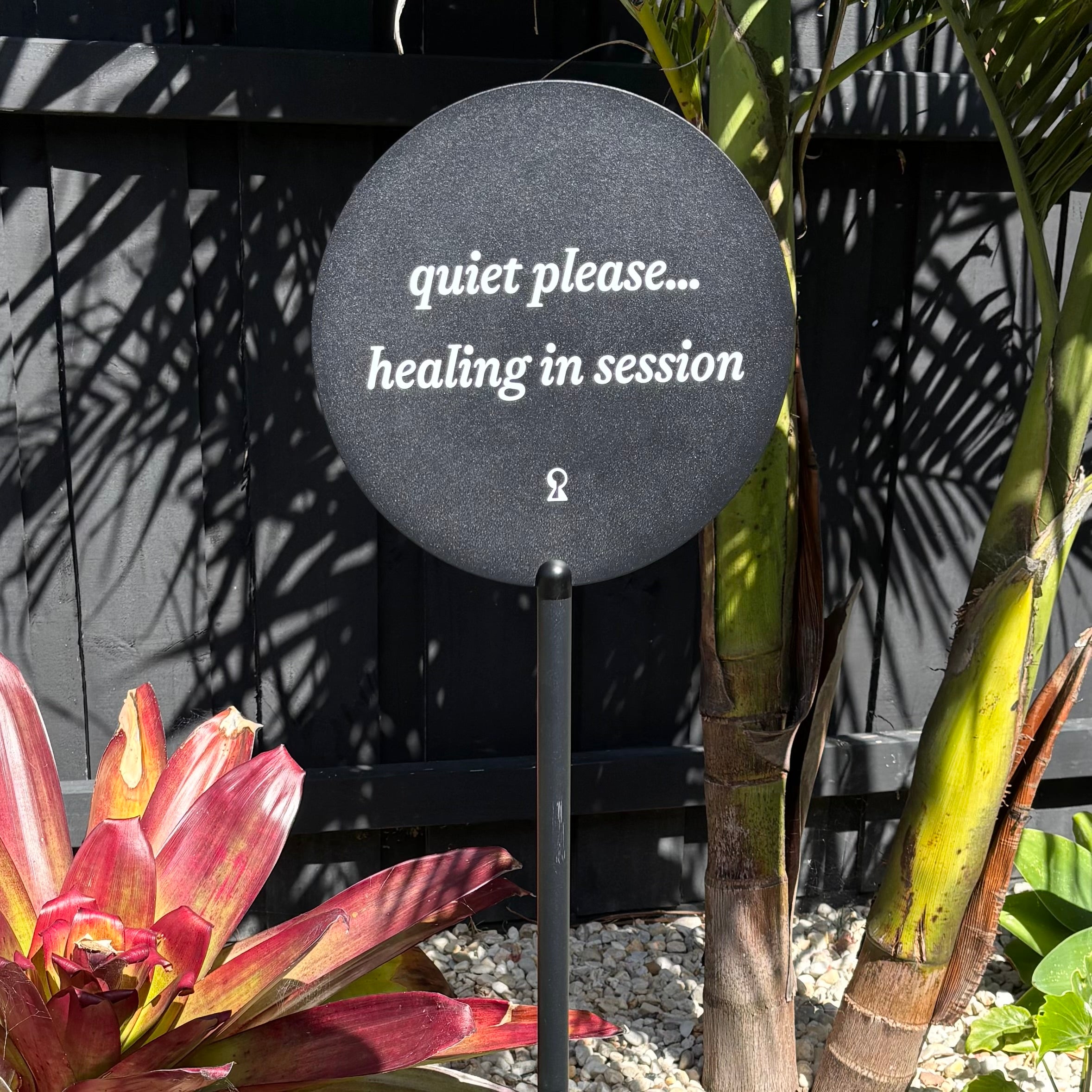 Black round freestanding acrylic sign reading “quiet please… healing in session”, designed for beauty clinics, wellness studios or treatment rooms, set outdoors among plants for a calm, professional aesthetic.