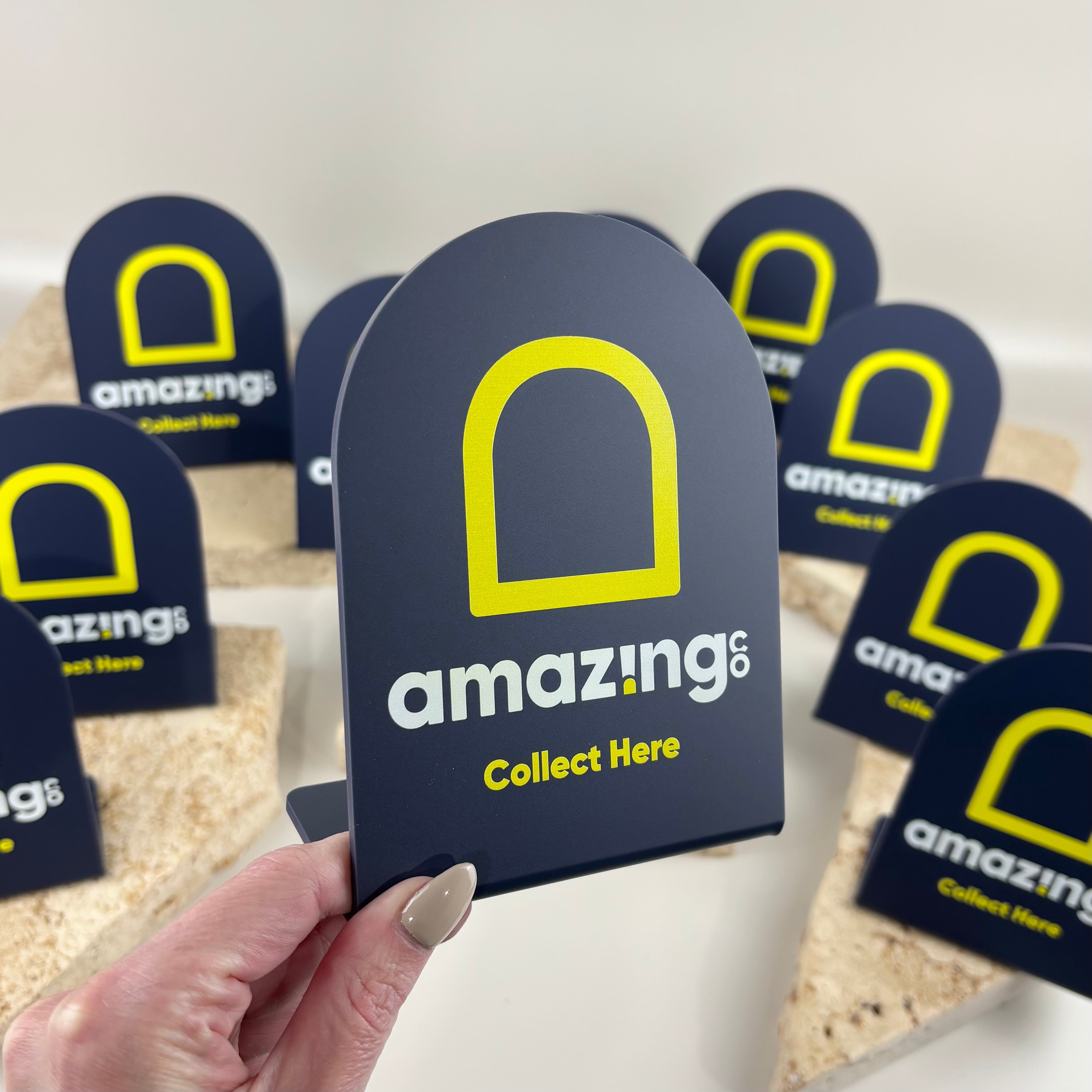 Custom UV printed acrylic collection counter signs with navy arch design and AmazingCo branding, featuring bright yellow and white Collect Here text.