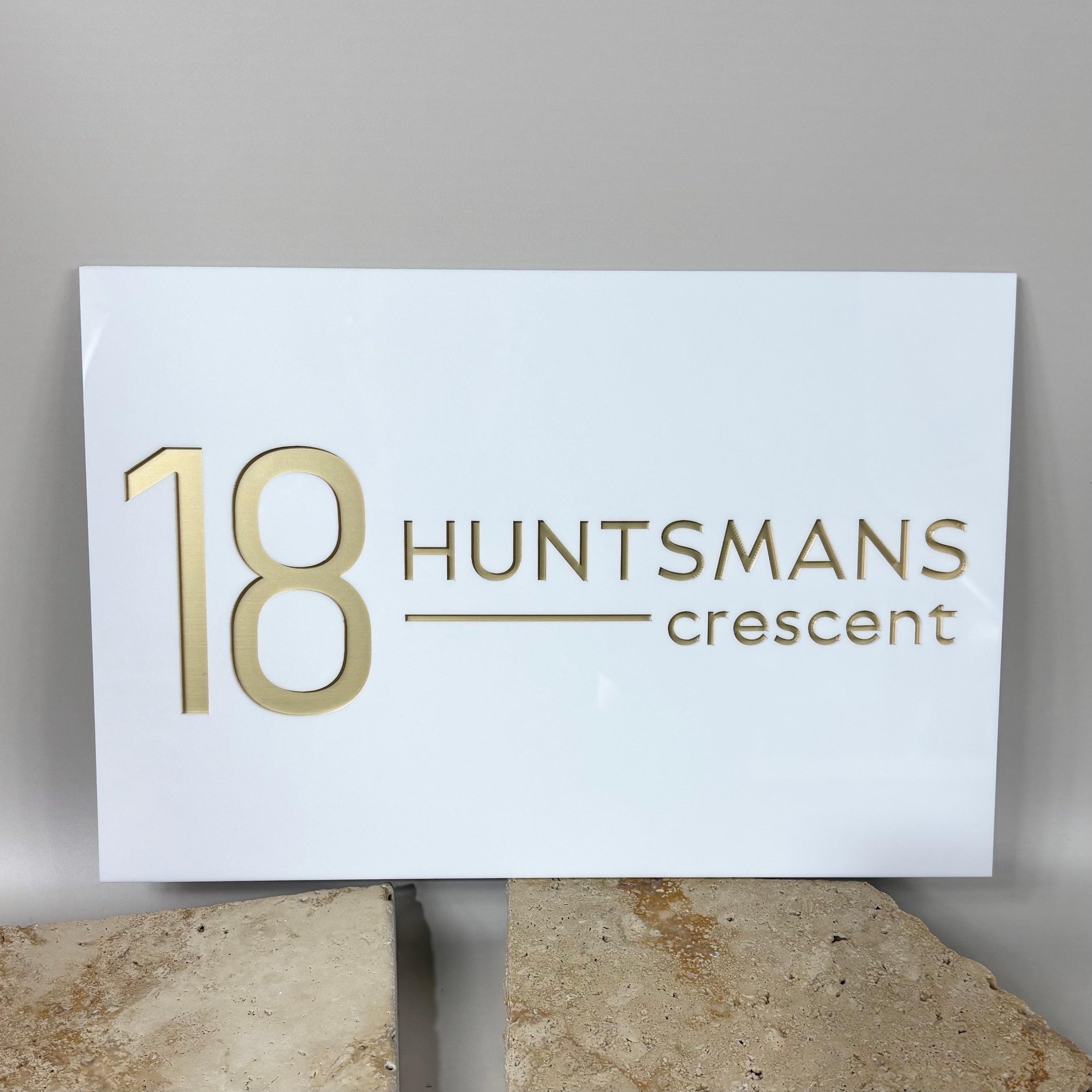 Custom white acrylic house sign with gold lettering, featuring the address “18 Huntsmans Crescent”, displayed on neutral stone background 