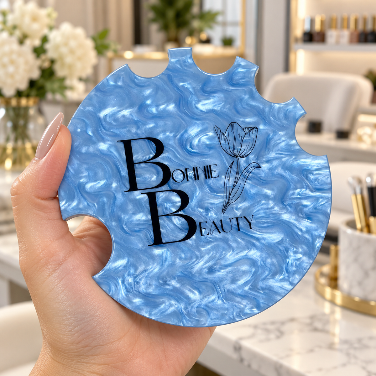 Custom blue pearl acrylic cup holder with Bonnie Beauty logo and floral design, held by a hand model with nude manicured nails in a modern beauty salon.